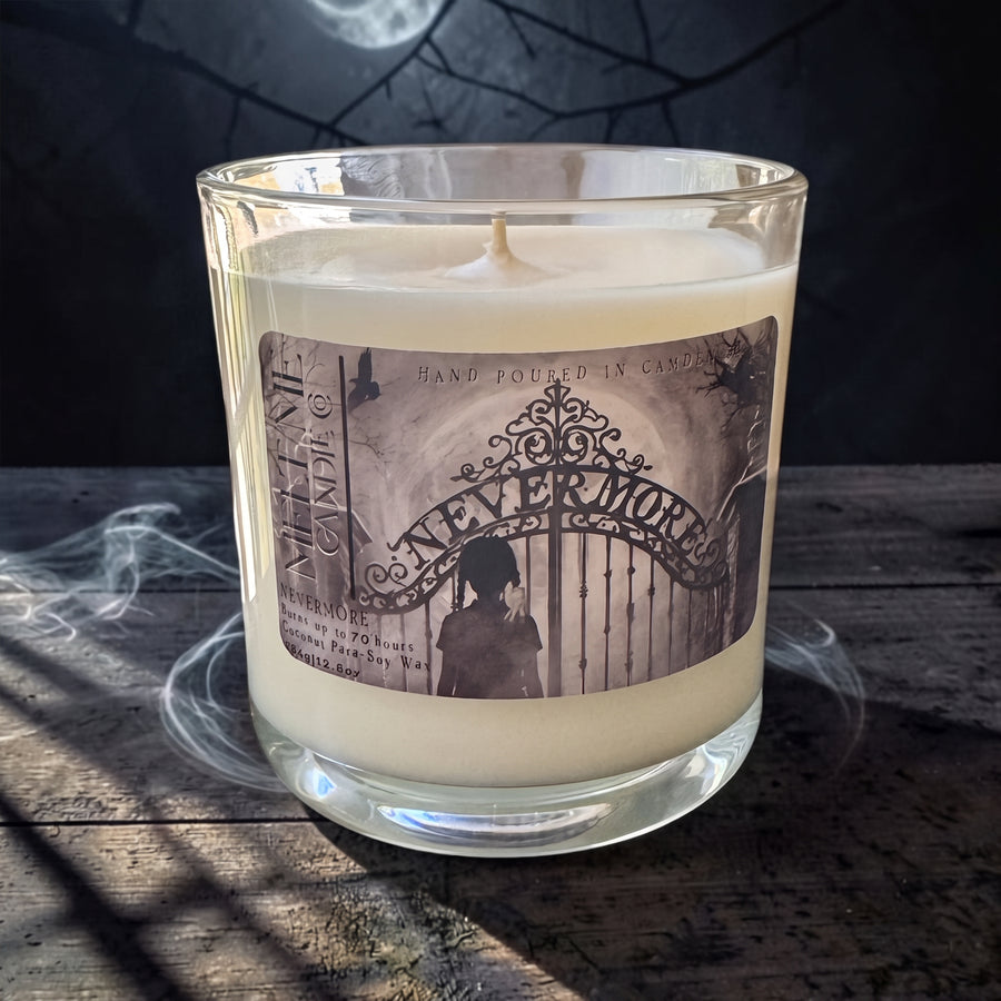 A clear glass jar candle filled with creamy off-white wax, featuring a vintage-style label that reads 'NEVER MOURN' with a sepia illustration of an ornate gate and a silhouette. The candle sits on a rustic dark wooden surface with dramatic shadows and a smoky, moody background.