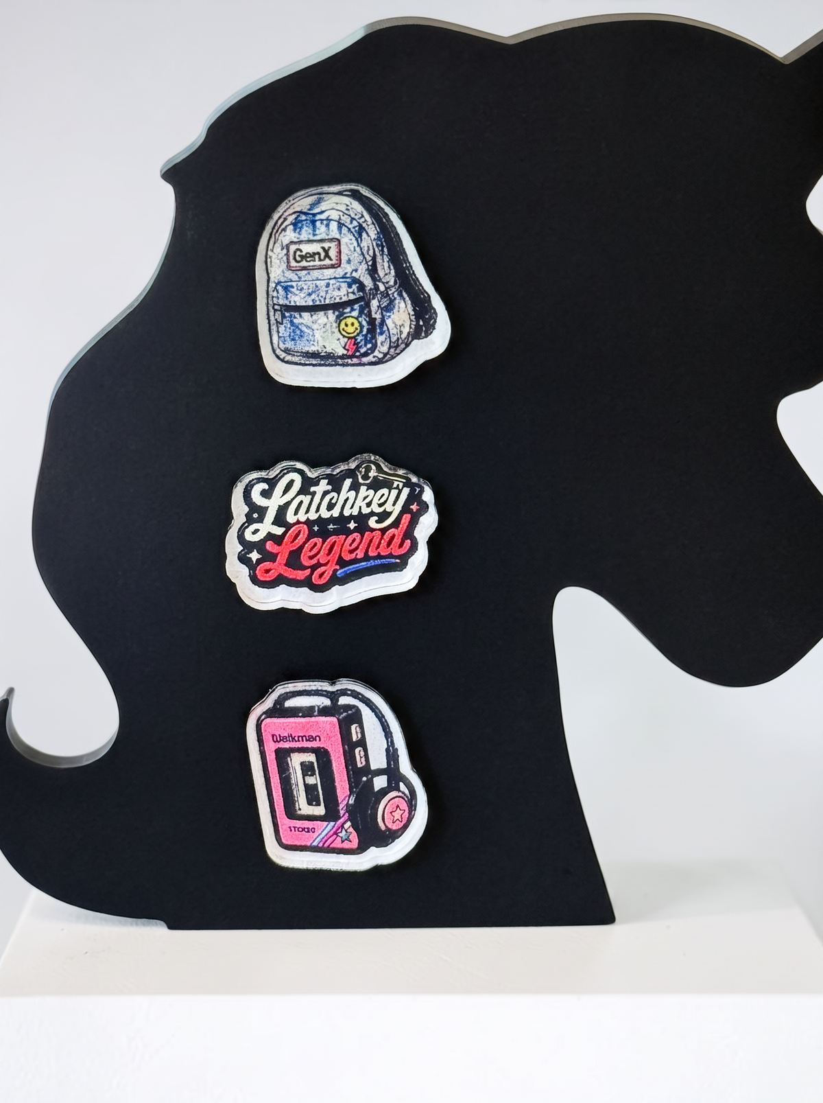 Three decorative earrings displayed on a black silhouette stand against a white background. The earrings feature a blue backpack, a pink and white 'Latchkey Legend' script design, and a pink cassette tape player with headphones.