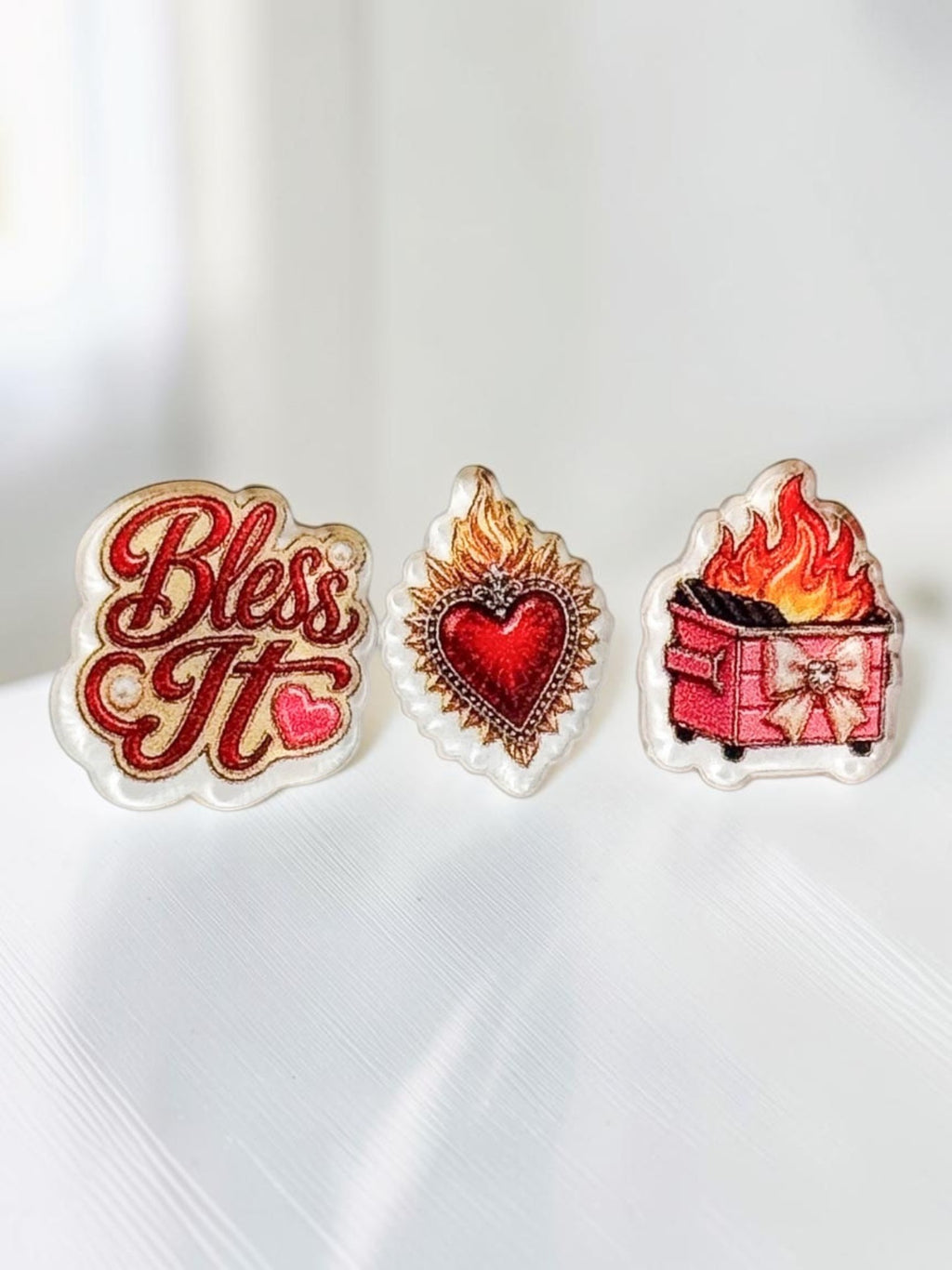 Three acrylic stud earrings displayed on a white surface: red script 'Bless It' with a bow, a sacred heart design, and a pink dumpster fire with flames and a bow.
