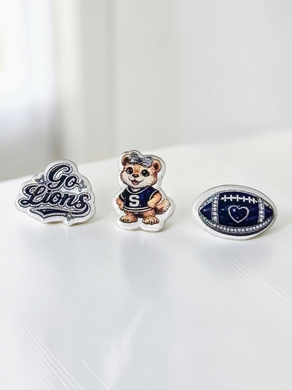 Three blue-and-white acrylic stud earrings displayed on a white surface: 'Go Lions' script, a lion mascot in a navy uniform, and a blue football.
