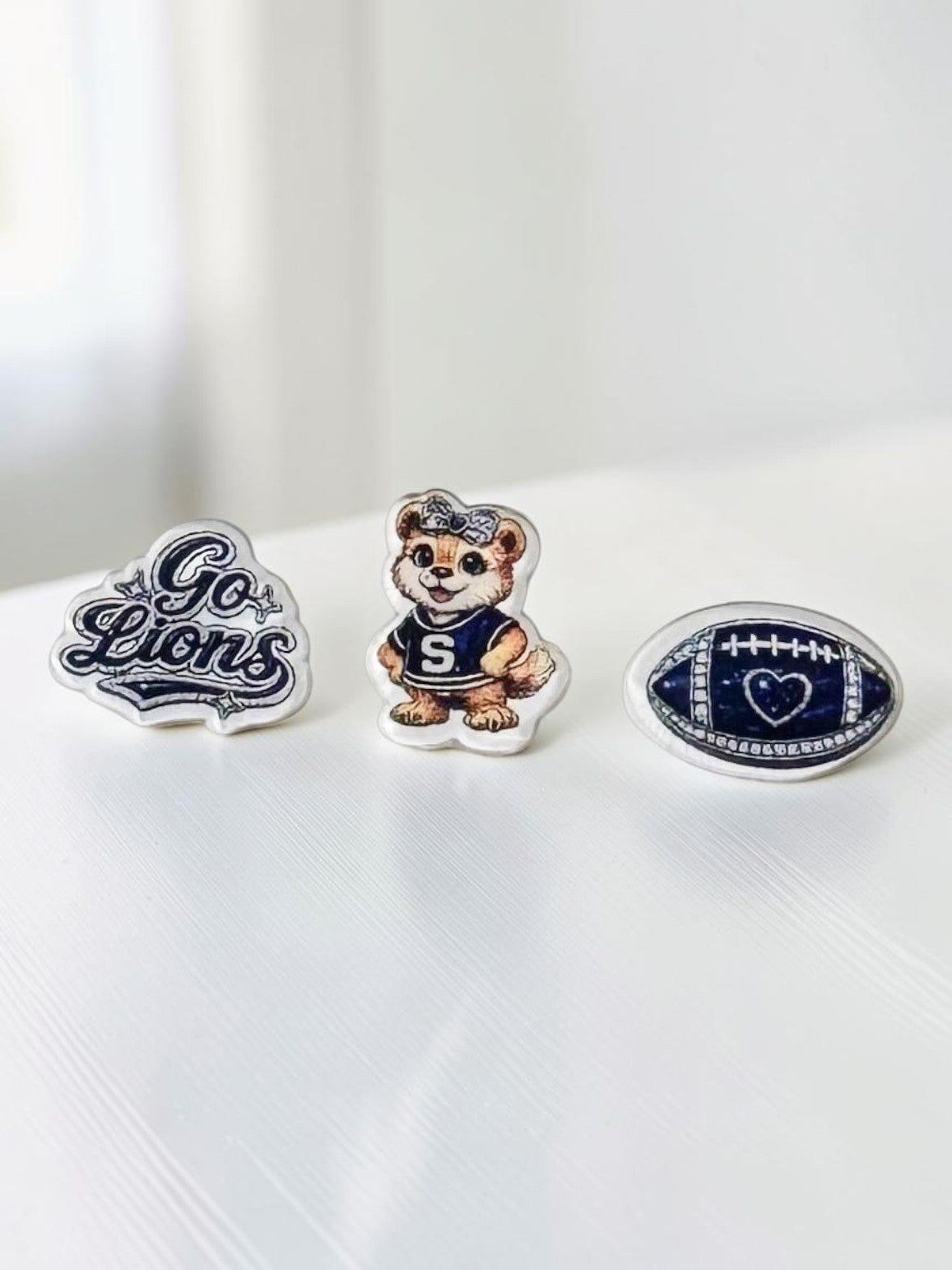 Three blue-and-white acrylic stud earrings displayed on a white surface: 'Go Lions' script, a lion mascot in a navy uniform, and a blue football.
