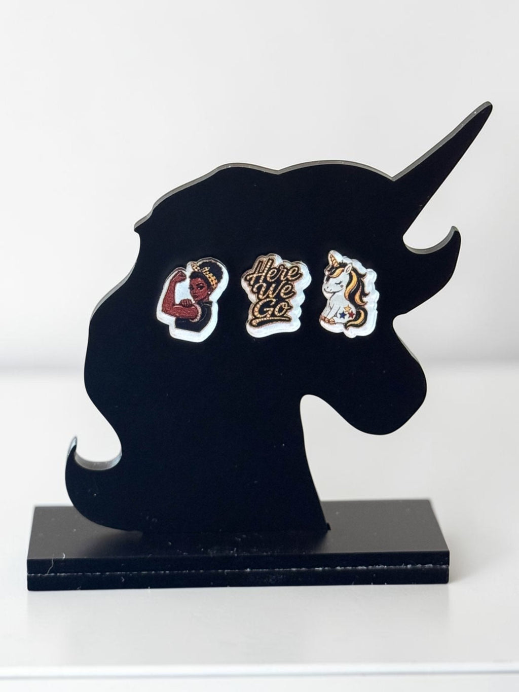 Three black-and-yellow acrylic stud earrings displayed on a black unicorn-shaped stand against a white background: a dark-skin Rosie-inspired flexing woman, a 'Here We Go' script design, and a white unicorn.