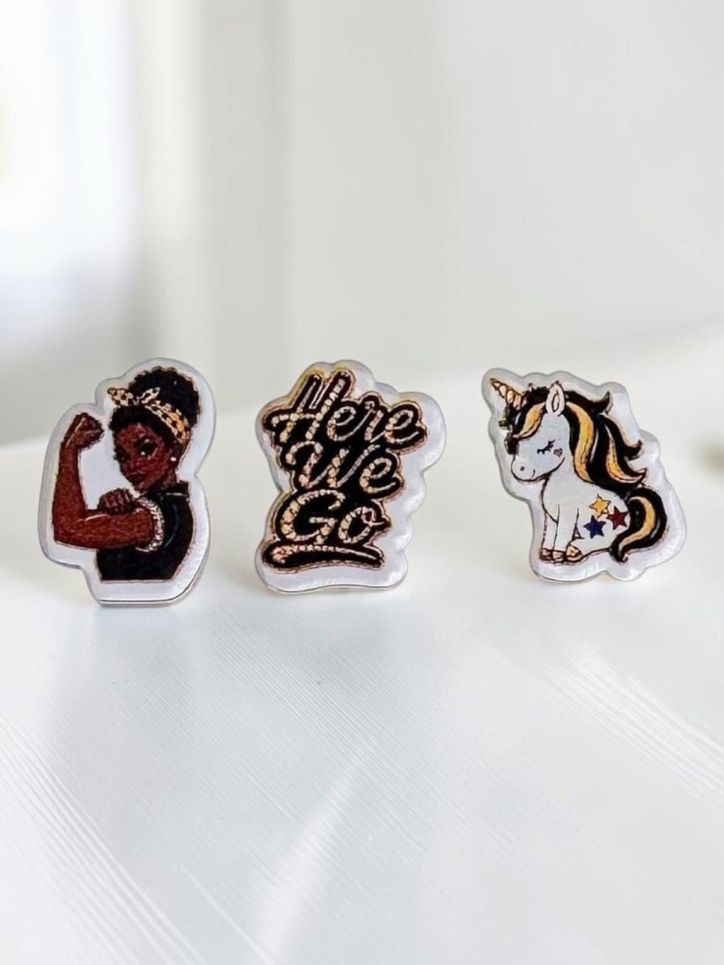 Three black-and-yellow acrylic stud earrings displayed on a white surface: a dark-skin Rosie-inspired flexing woman, a 'Here We Go' script design, and a unicorn with gold and pink details.