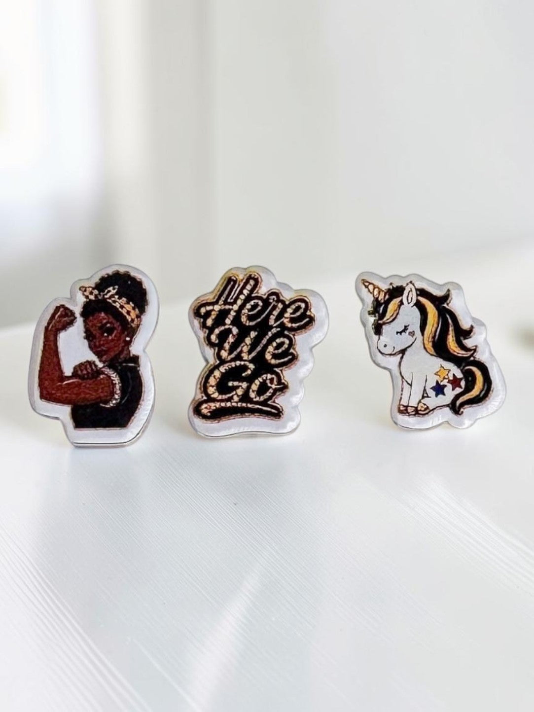 Three black-and-yellow acrylic stud earrings displayed on a white surface: a dark-skin Rosie-inspired flexing woman, a 'Here We Go' script design, and a unicorn with gold and pink details.