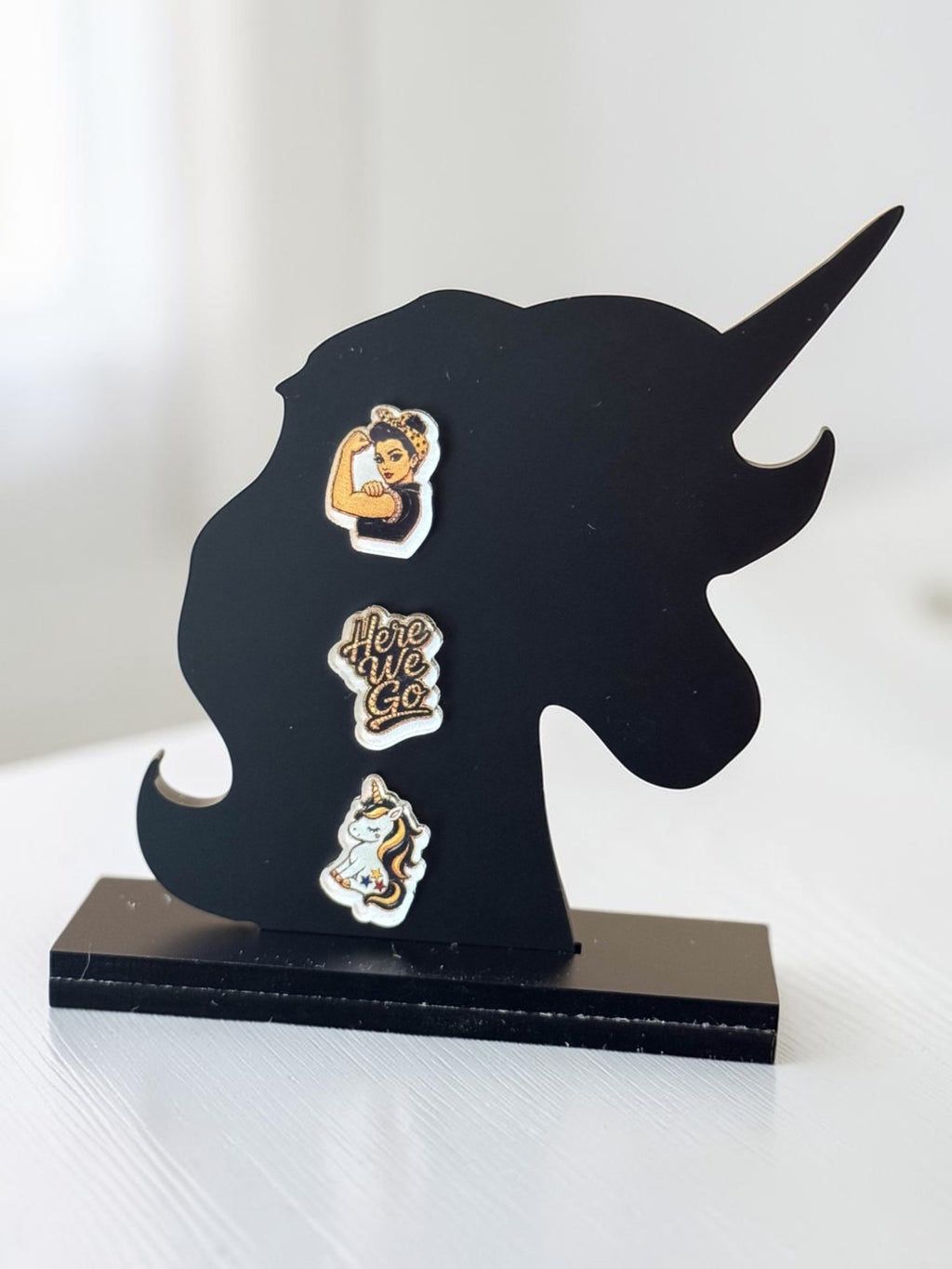 Three black-and-yellow acrylic stud earrings displayed on a black unicorn-shaped stand: a fair-skin Rosie-inspired flexing woman, a 'Here We Go' script design, and a unicorn.