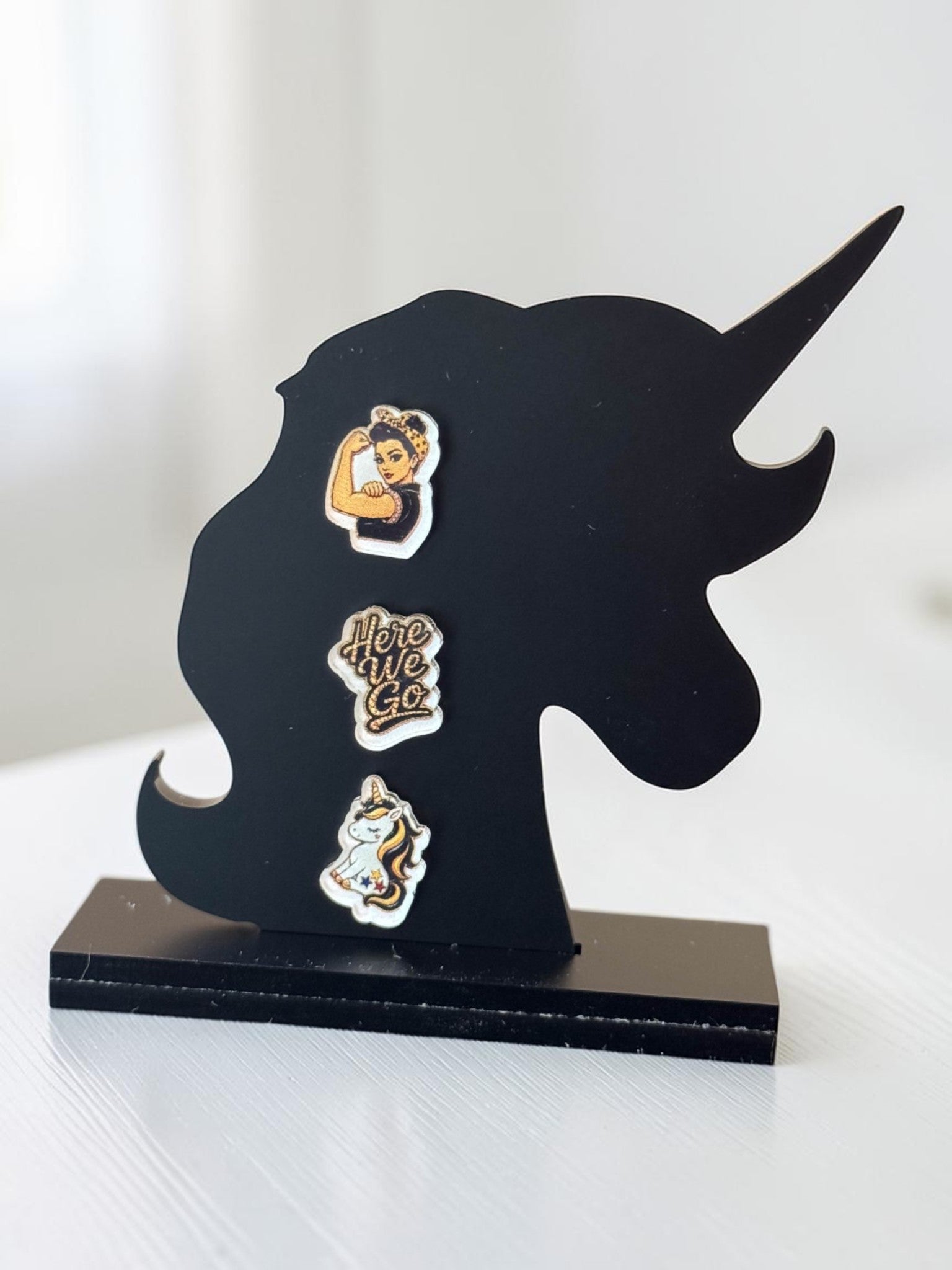 Three black-and-yellow acrylic stud earrings displayed on a black unicorn-shaped stand: a fair-skin Rosie-inspired flexing woman, a 'Here We Go' script design, and a unicorn.