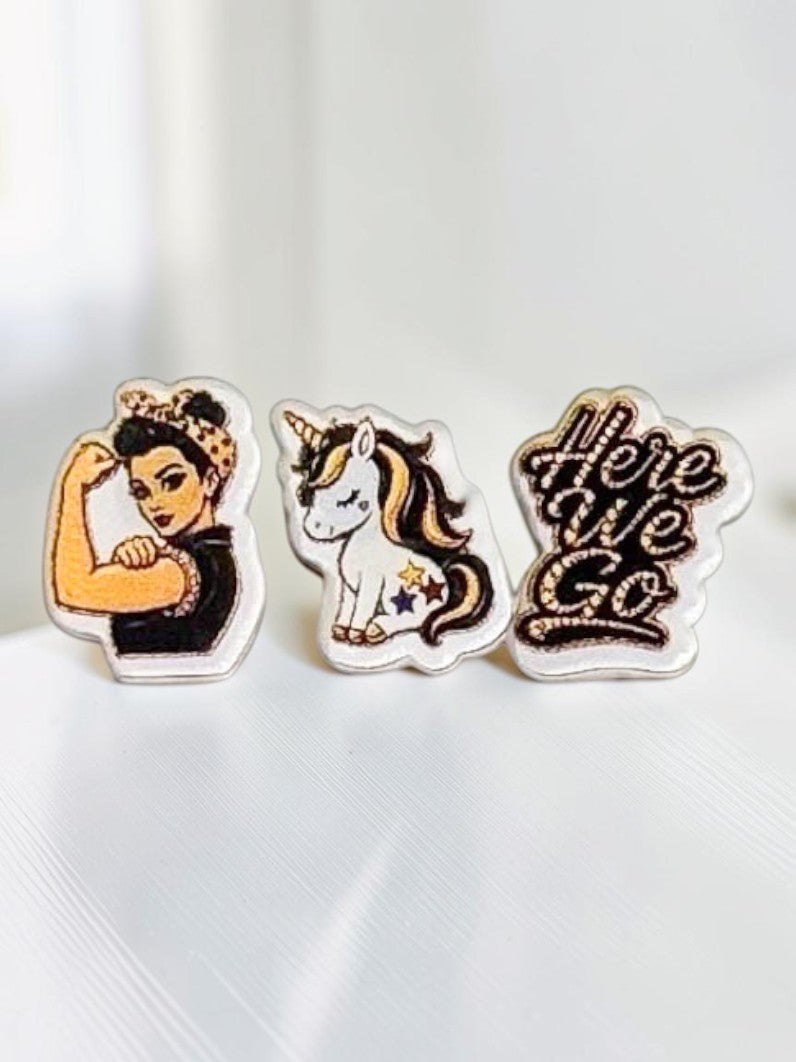 Three black-and-yellow acrylic stud earrings displayed on a white surface: a fair-skin Rosie-inspired flexing woman, a 'Here We Go' script design, and a unicorn.