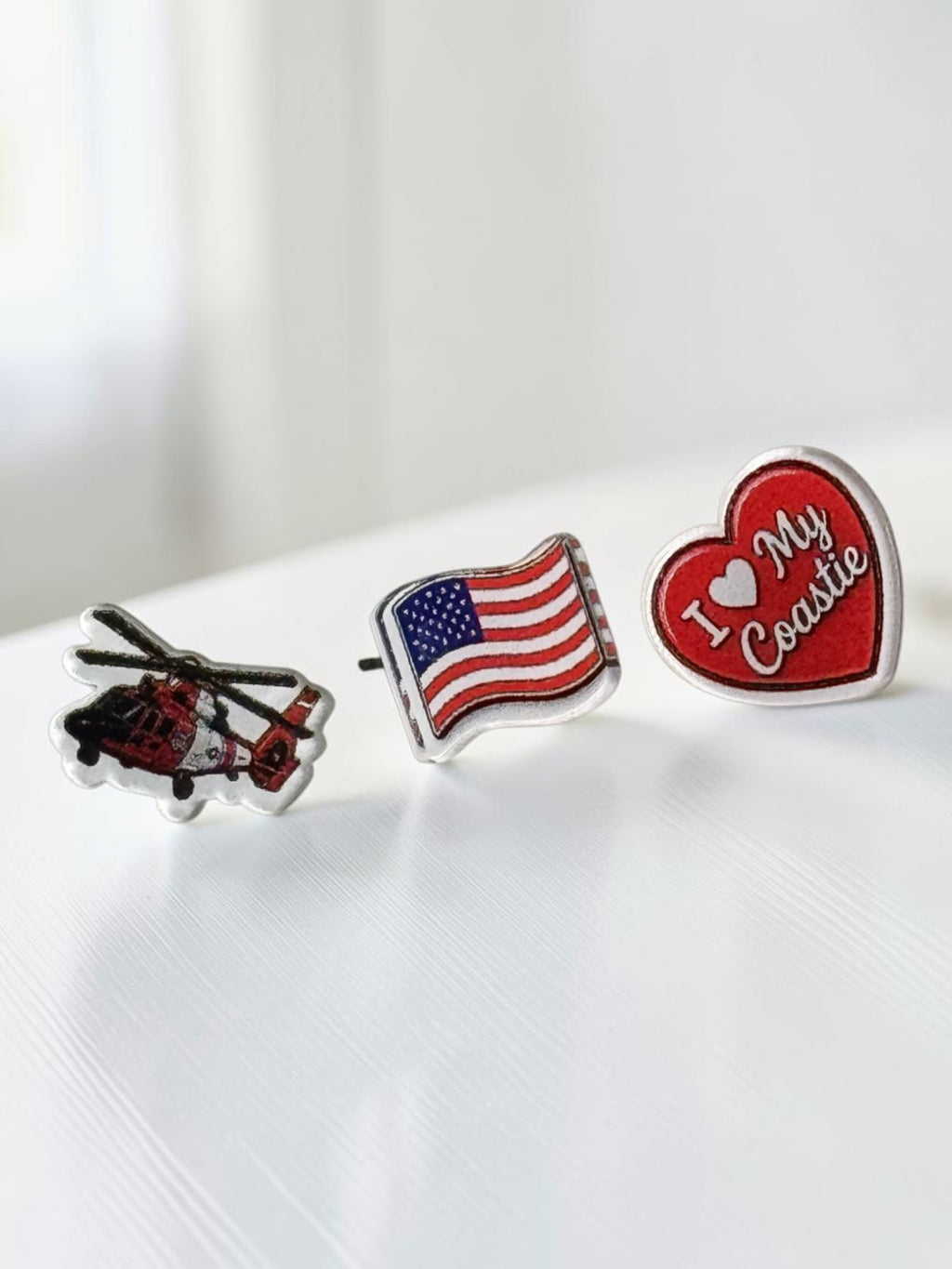 Three small enamel pins on a white surface: a helicopter pin, an American flag pin, and a red heart pin that says "I ❤️ My Coastie" in white script.