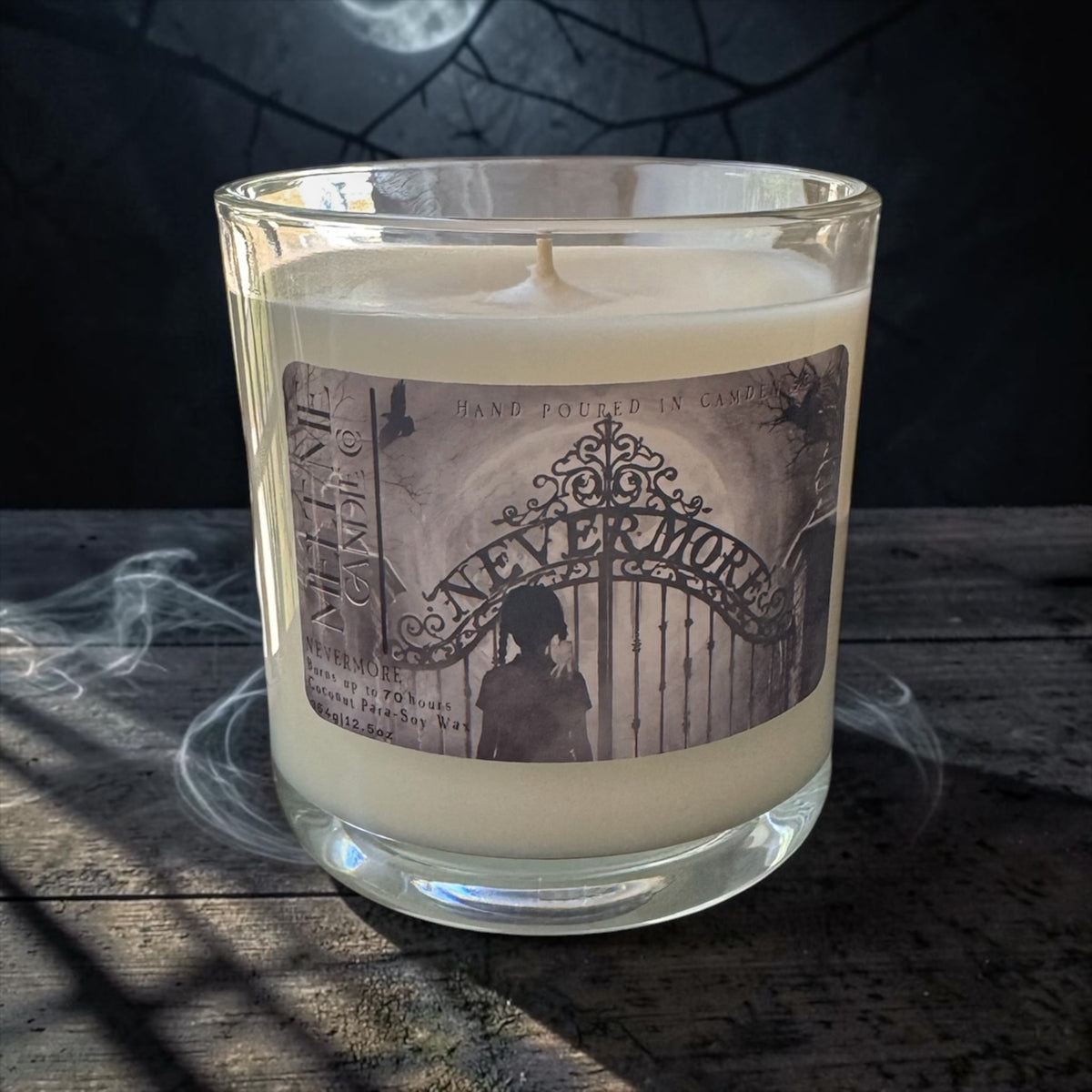 A clear glass jar candle filled with creamy off-white wax, featuring a vintage-style label that reads 'NEVER MOURN' with a sepia illustration of an ornate gate and a silhouette. The candle sits on a rustic dark wooden surface with dramatic shadows and a smoky, moody background.