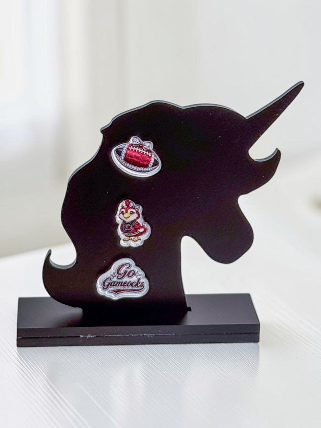 A black unicorn-shaped display stand on a rectangular base holding three garnet-and-black acrylic stud earrings: a garnet football, a 'Go Fight Win' design, and a bird mascot, photographed on a white surface against a softly blurred background.