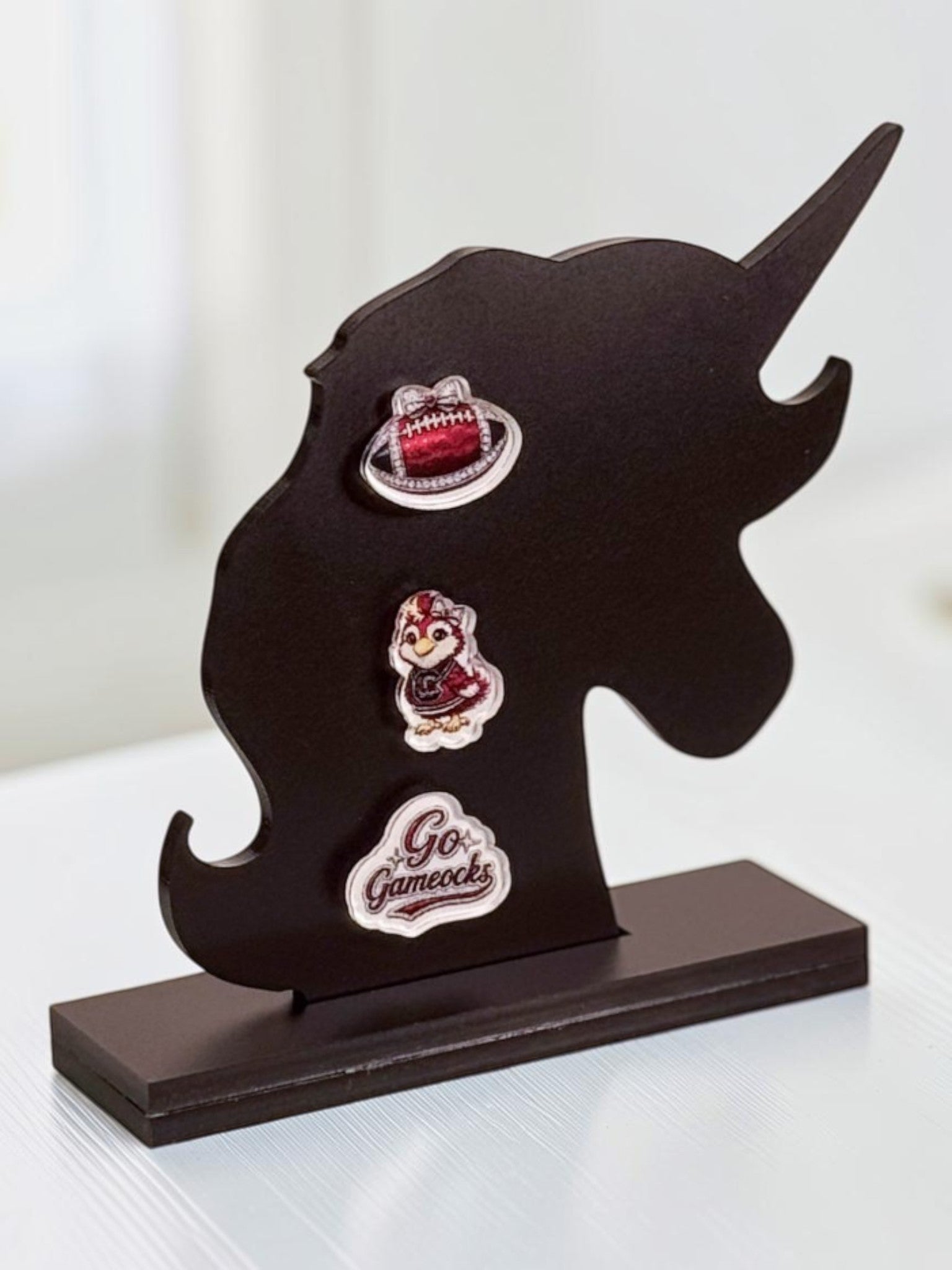 A unicorn-shaped tabletop display stand holding three garnet-and-black acrylic stud earrings: a garnet football, a 'Go Fight Win' design, and a bird mascot, photographed against a softly blurred indoor background.