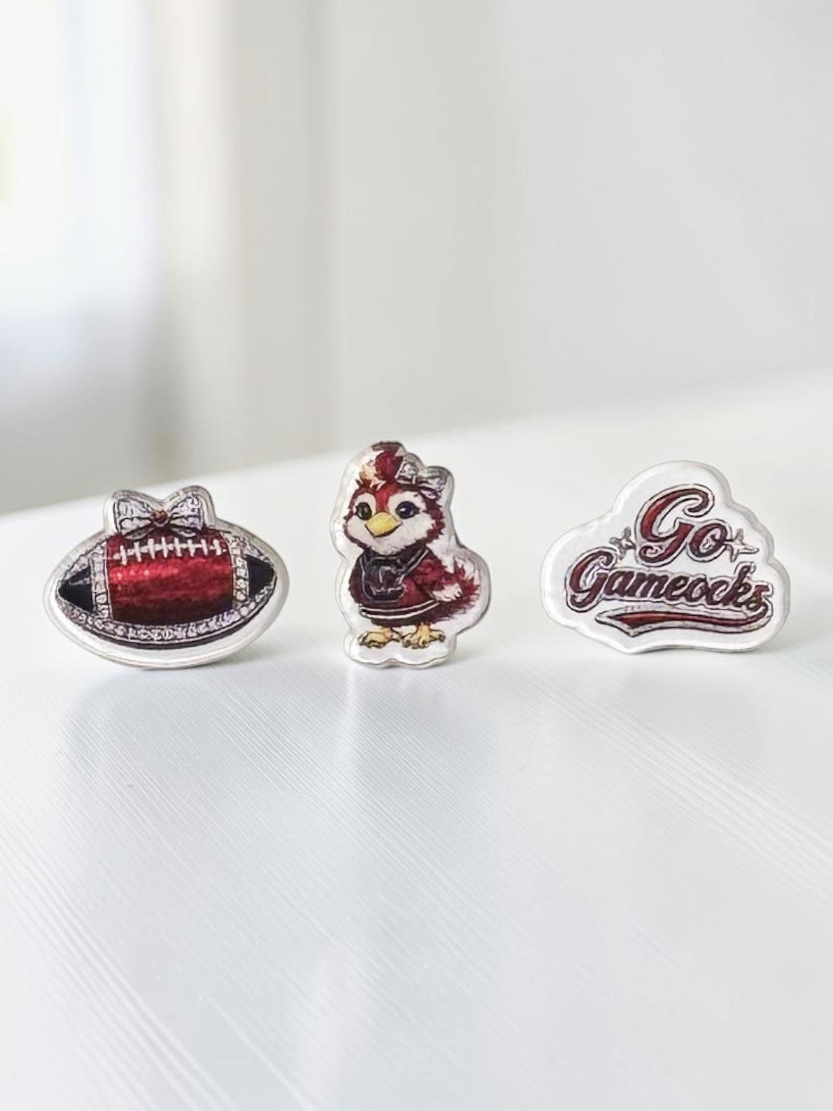 Three garnet-and-black acrylic stud earrings displayed on a white surface: a garnet football, a 'Go Fight Win' script design, and a bird mascot in a garnet-and-white uniform.
