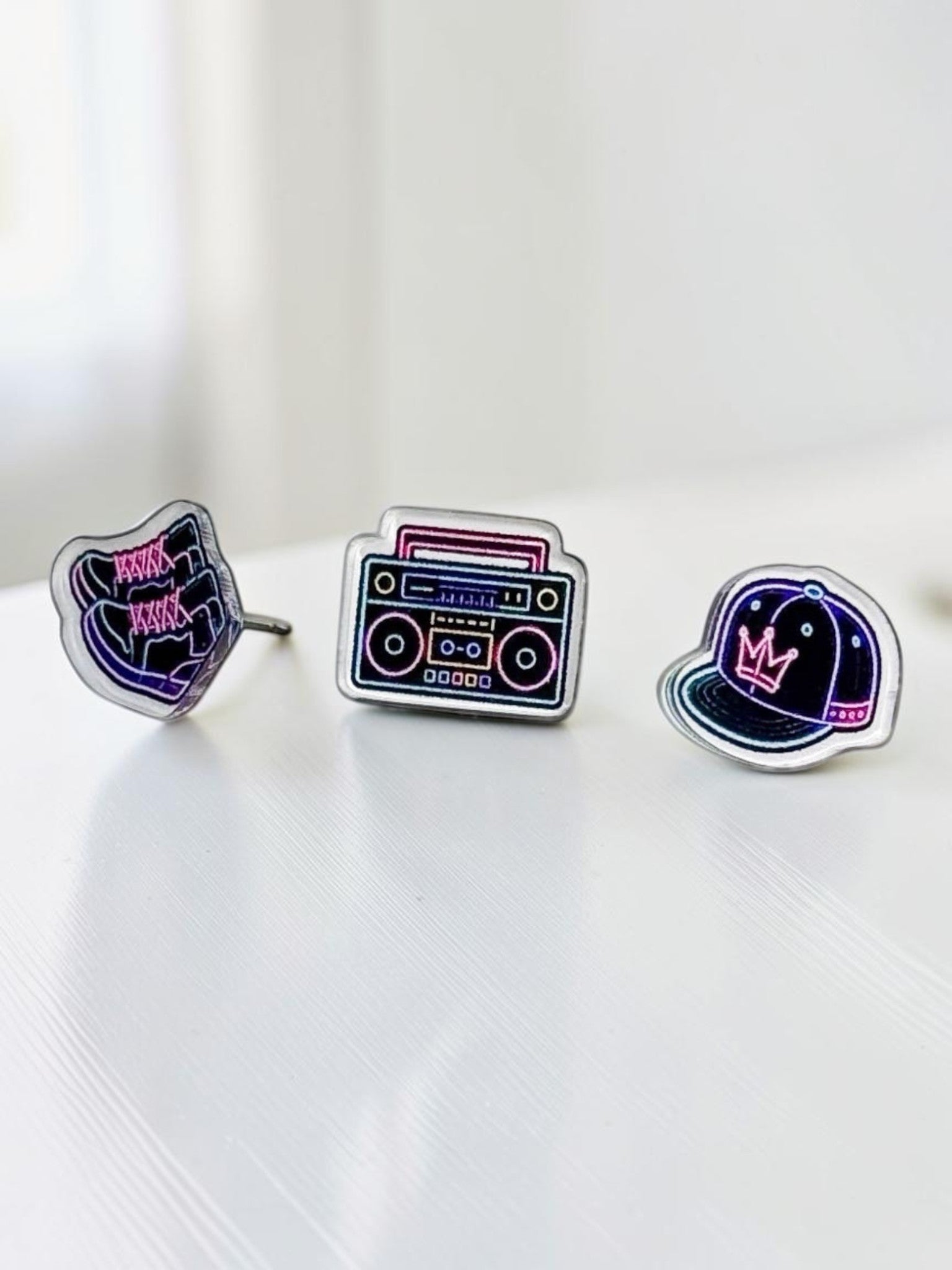 Three neon acrylic stud earrings displayed on a white surface: bright sneakers, a colorful streetwear hat, and a retro boombox.