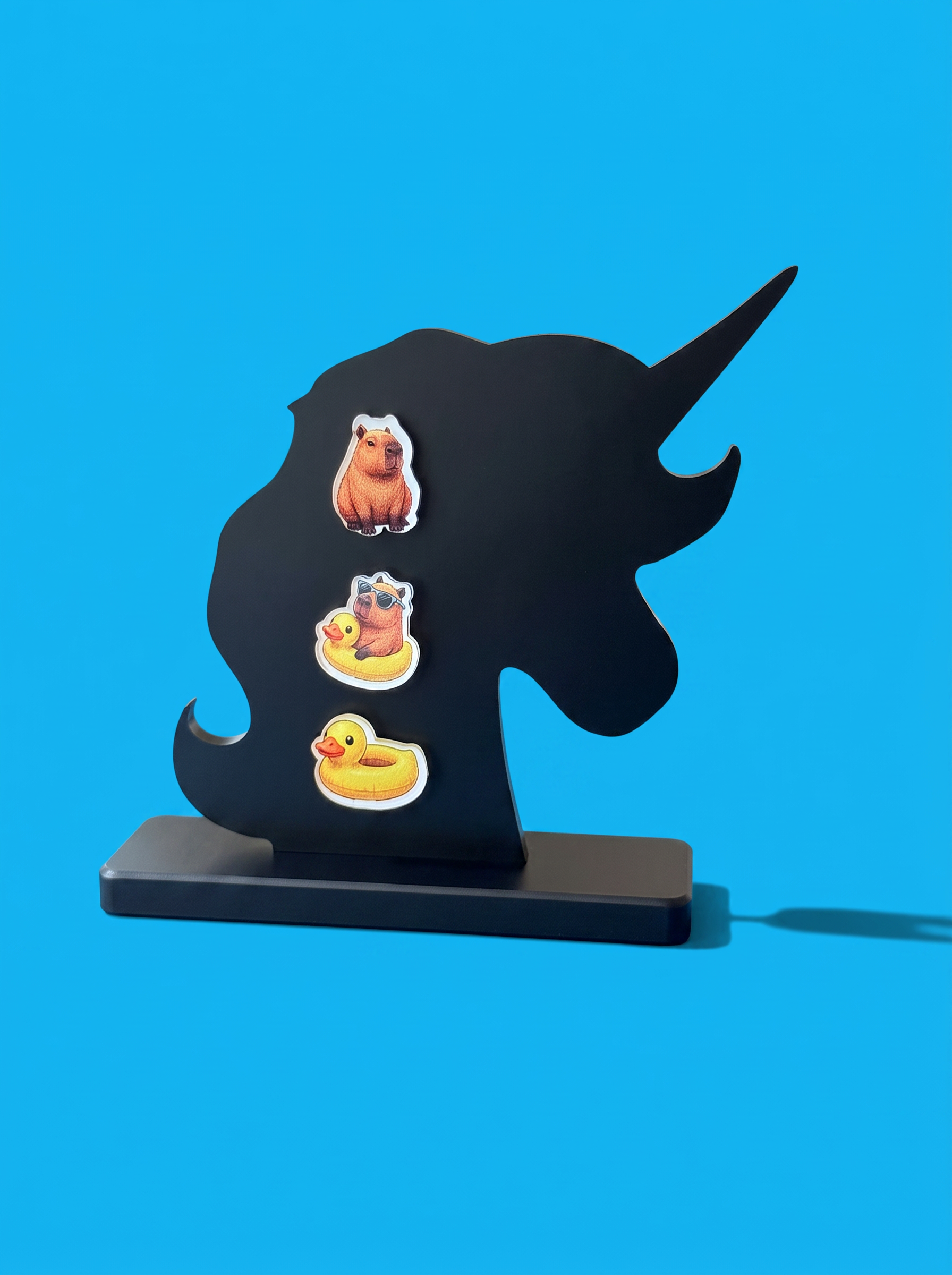 A black unicorn-shaped display stand on a matching base, featuring a capybara, a capybara in a ducky raft and a ducky raft photographed against a bright blue background.