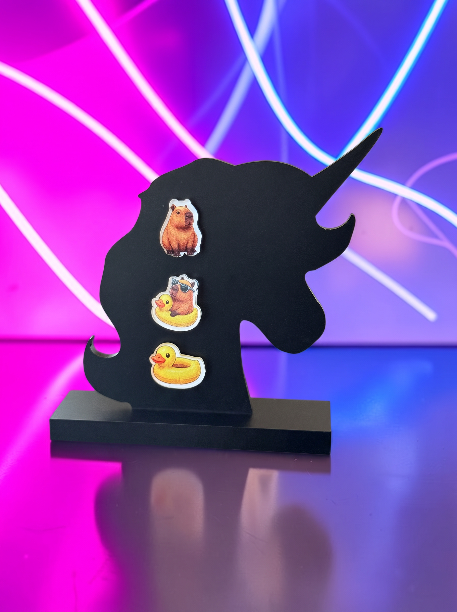 A three earring set featuring a  capybara, a capybara in a ducky raft and a ducky raft displayed on a black base against a neon pink and blue light background.
