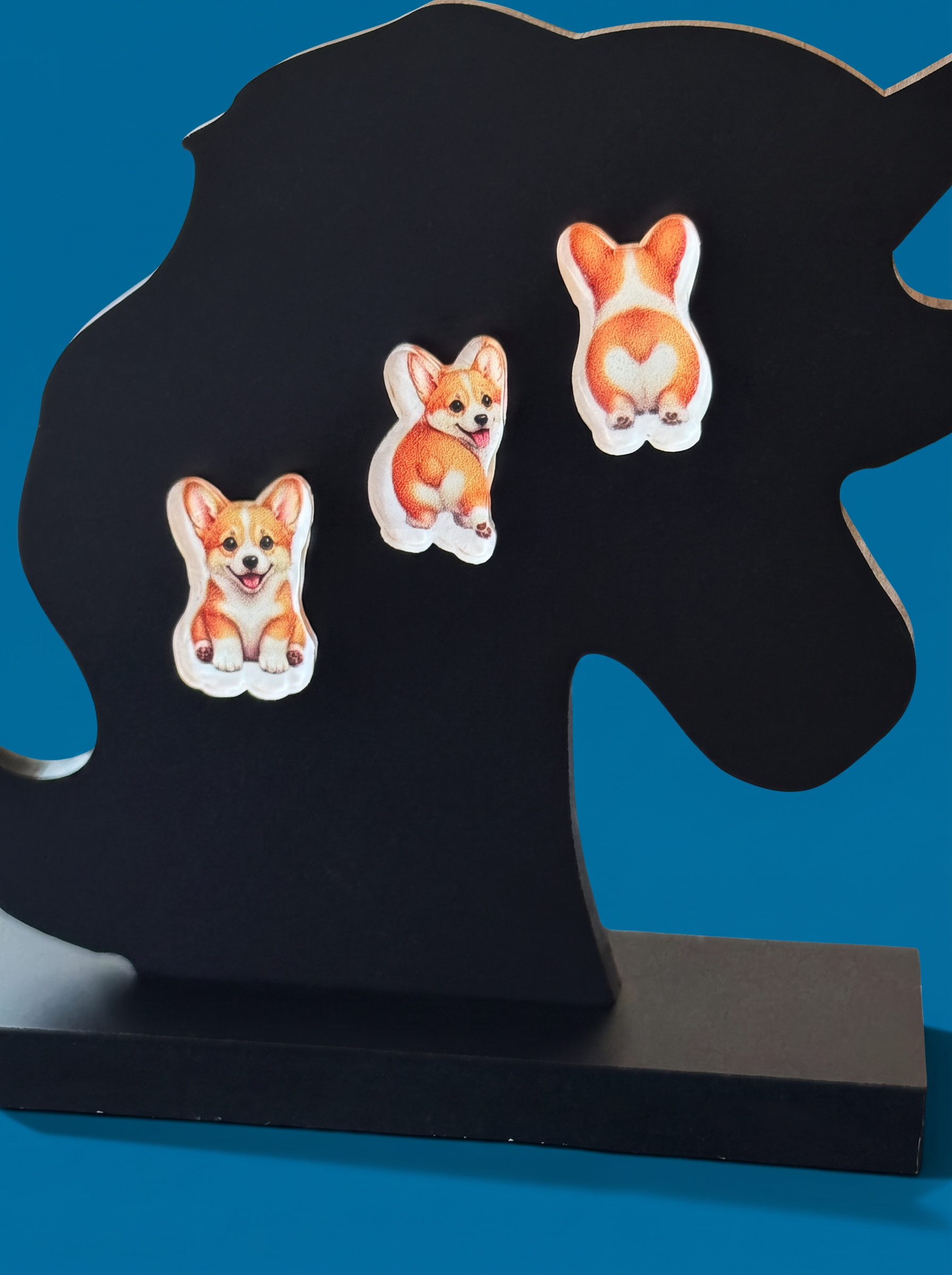 Three small earrings with illustrated corgi faces and backsides are arranged on a black silhouette stand against a bright blue background.