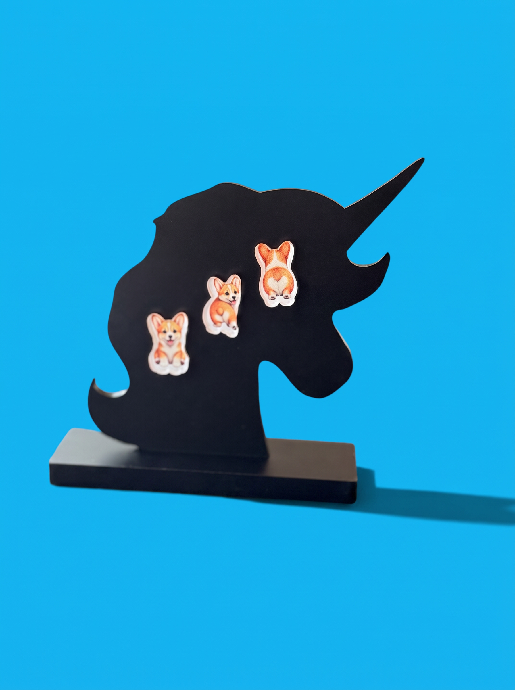 Corgi dog earrings with orange and white coloring are displayed on a black unicorn silhouette stand against a bright blue background.
