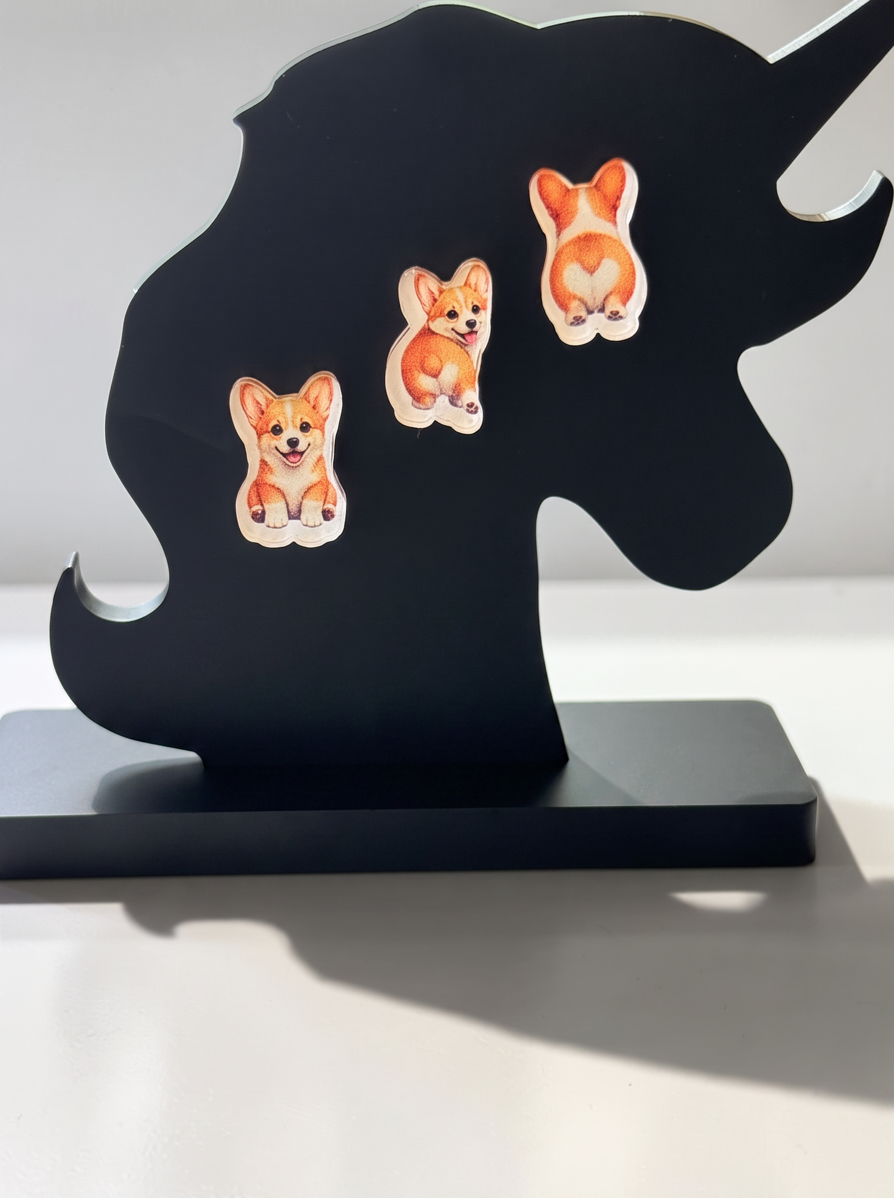 Three decorative earrings displayed on a black unicorn-shaped stand against a light gray background. The earrings include a corgi sitting, a corgi from behind and a corgi looking over their shoulder showing off that fluffy tush.