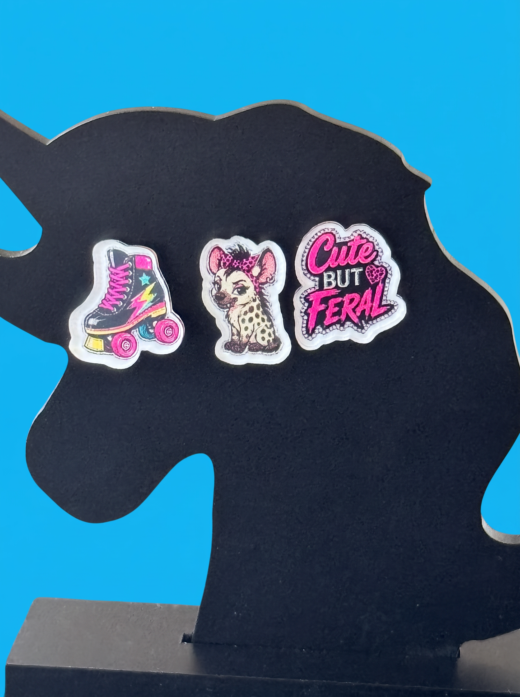 Three decorative earrings displayed on a black unicorn-shaped stand against a bright blue background. The earrings include a pink roller skate, a spotted hyena with a hair bow, and the phrase 'Cute But Feral' in pink lettering