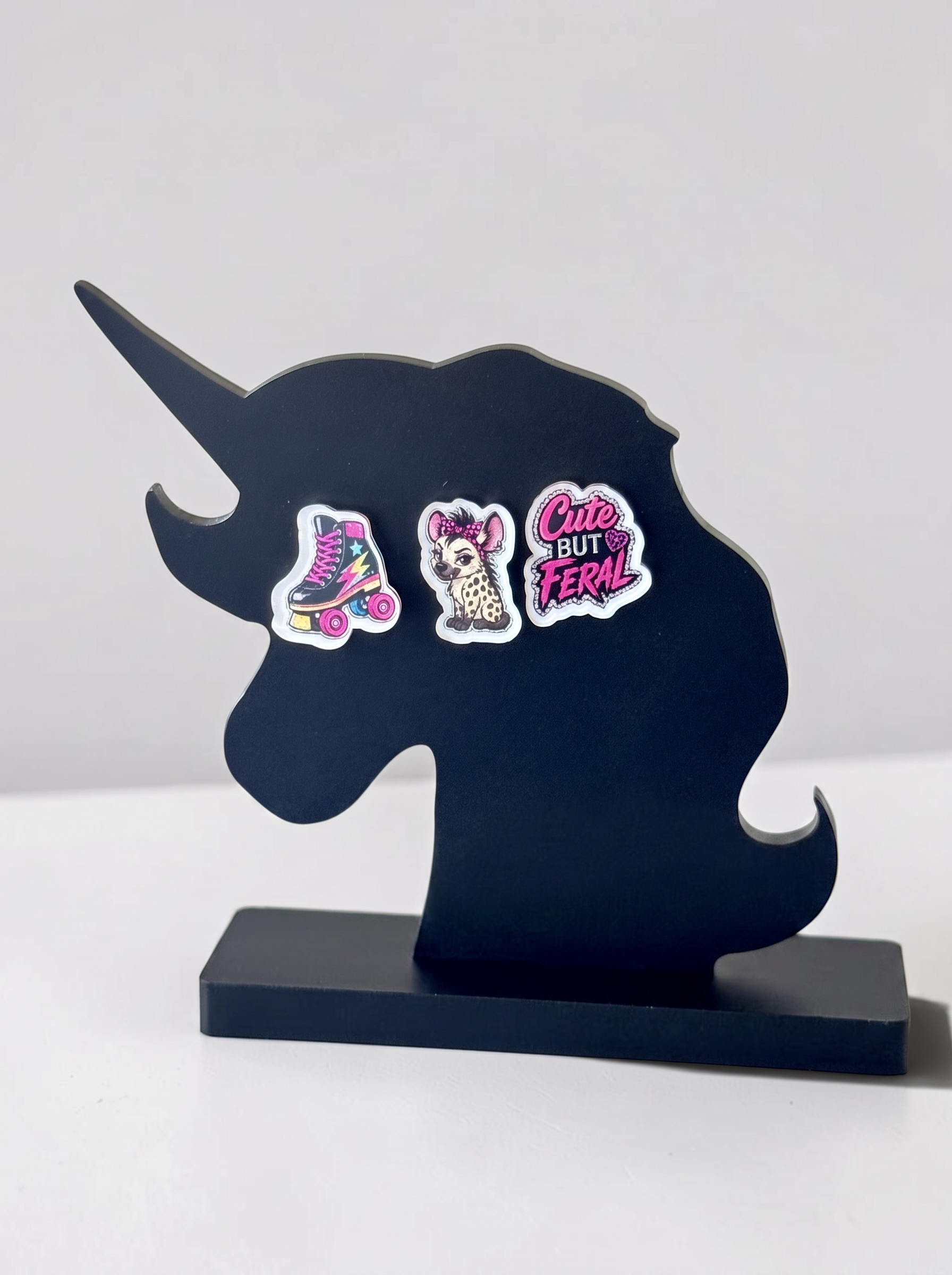 A black unicorn-shaped decorative stand or plaque on a matching rectangular base, against a white background. Three earrings include a pink roller skate, a spotted hyena with a hair bow, and the phrase 'Cute But Feral' in pink lettering. 