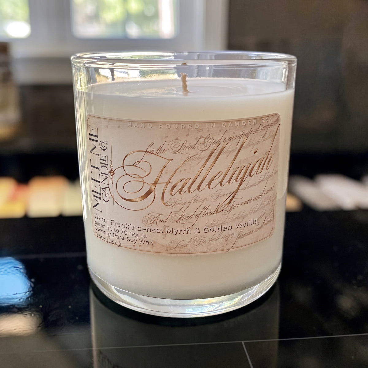 A clear glass jar candle filled with creamy off-white wax, featuring a decorative pink label with elegant script reading 'Hallelujah' and vertical text for 'The Melting Candle'. The candle is photographed on a glossy black surface with a dark, blurred background.