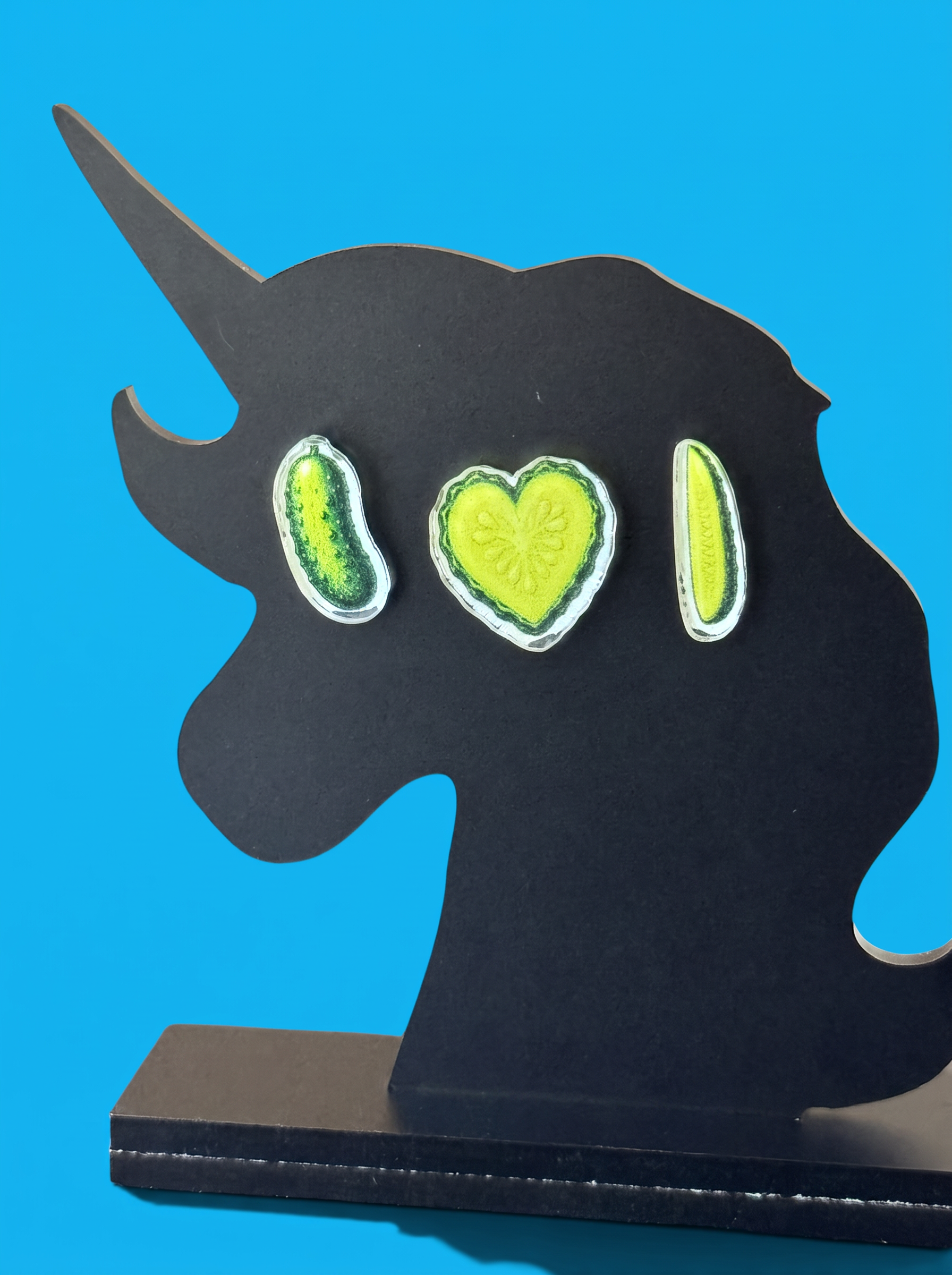 A black unicorn-shaped decorative plaque or sign with three pickle-themed earring designs, displayed on a black base against a bright blue background.