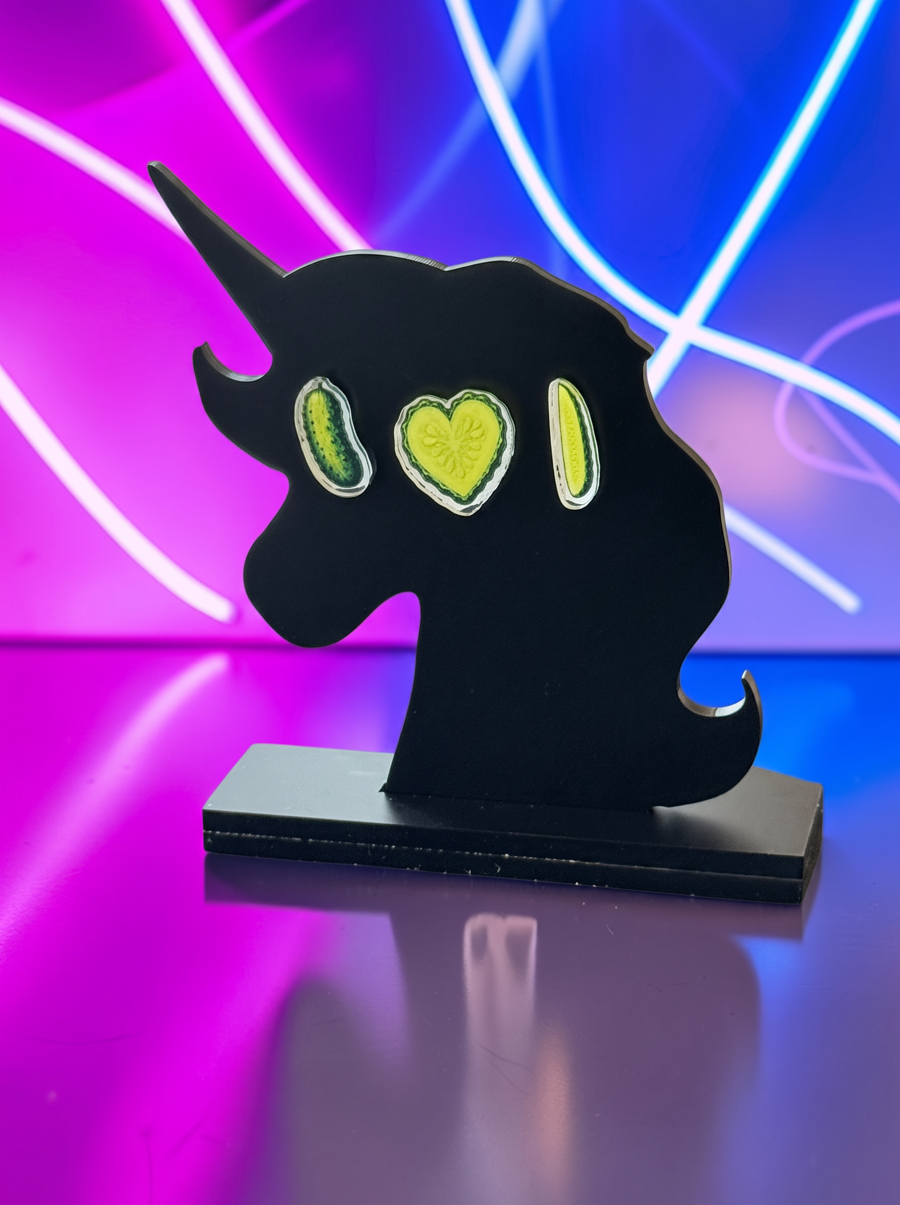 A black unicorn-shaped decorative unicorn with three lime-green pickle earrings on a neon pink and blue light background.
