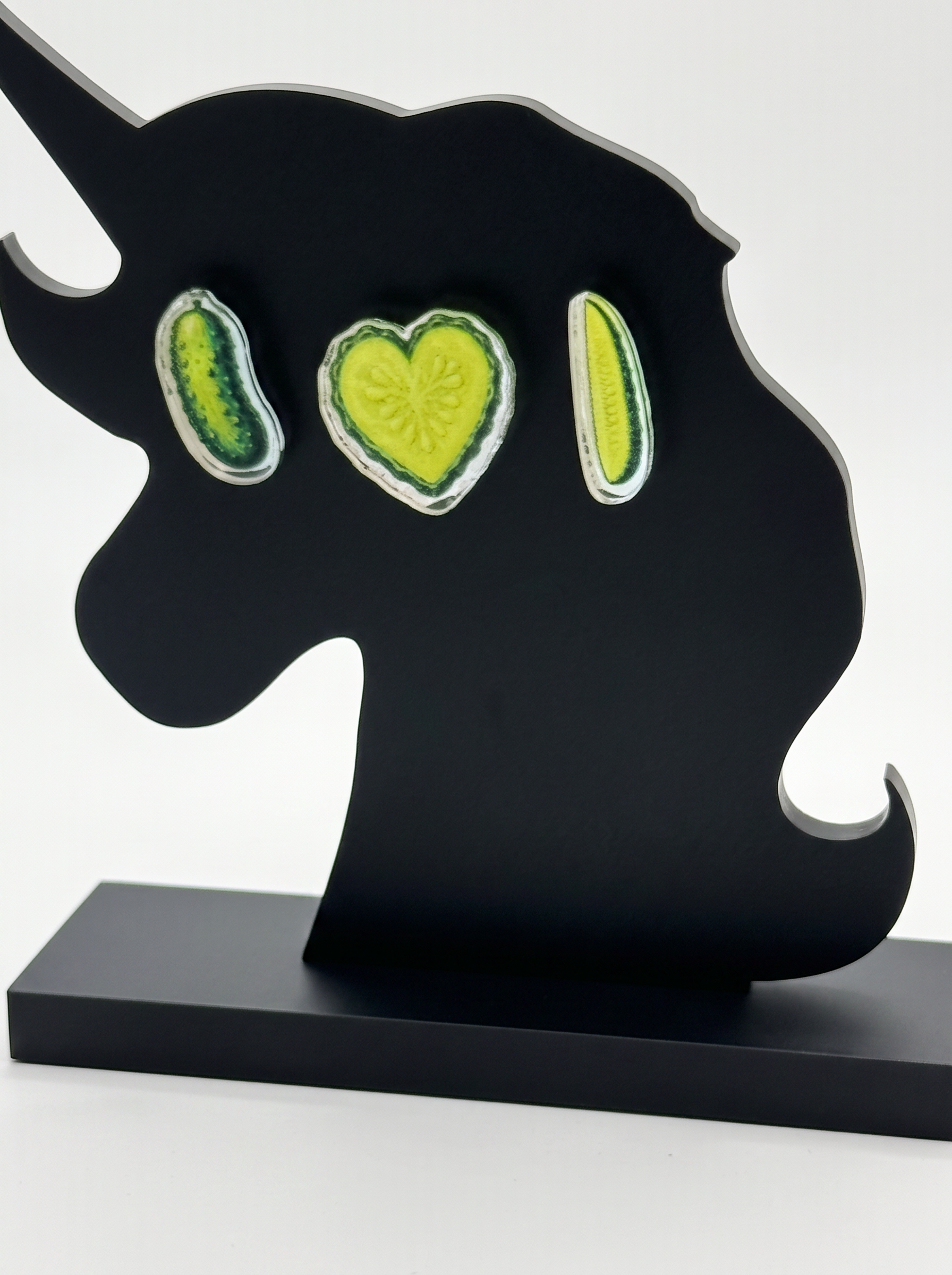 A black metal unicorn-shaped wall decor A black unicorn-shaped decorative earring stand with three pickle shaped earrings. A whole pickle, a heart-shaped pickle slice, and a spear, displayed on a black unicorn silhouette against a white background