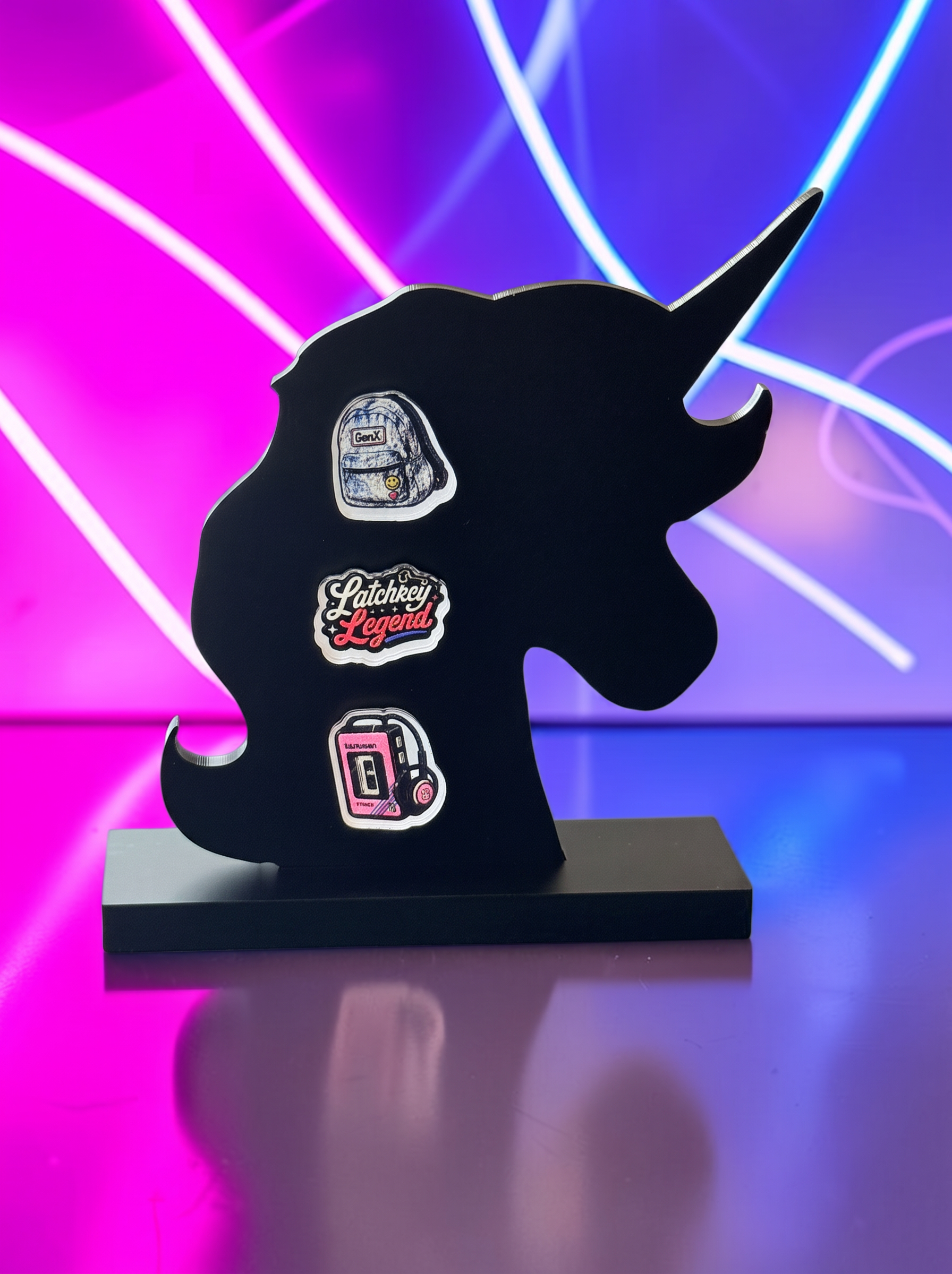 Three decorative earrings displayed on a black silhouette stand against a blue and pink neon background. The earrings feature a blue backpack, a pink and white 'Latchkey Legend' script design, and a pink cassette tape player with headphones.