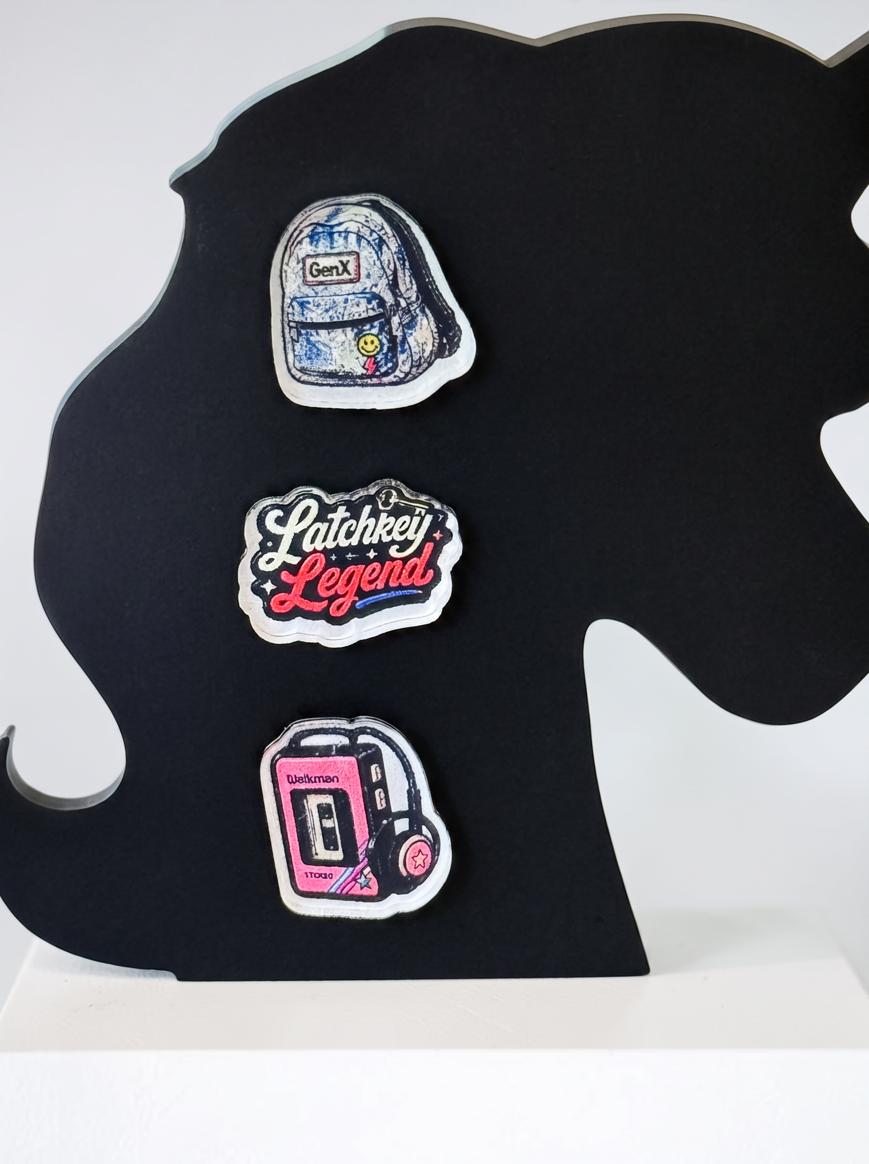 Three decorative earrings displayed on a black silhouette stand against a white background. The earrings feature a blue backpack, a pink and white 'Latchkey Legend' script design, and a pink cassette tape player with headphones.