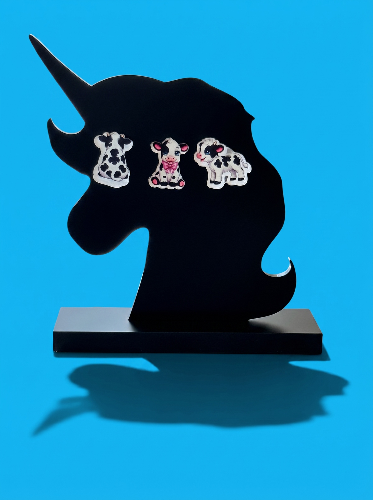 Three cow-shaped earrings displayed on a black unicorn head stand against a bright blue background.