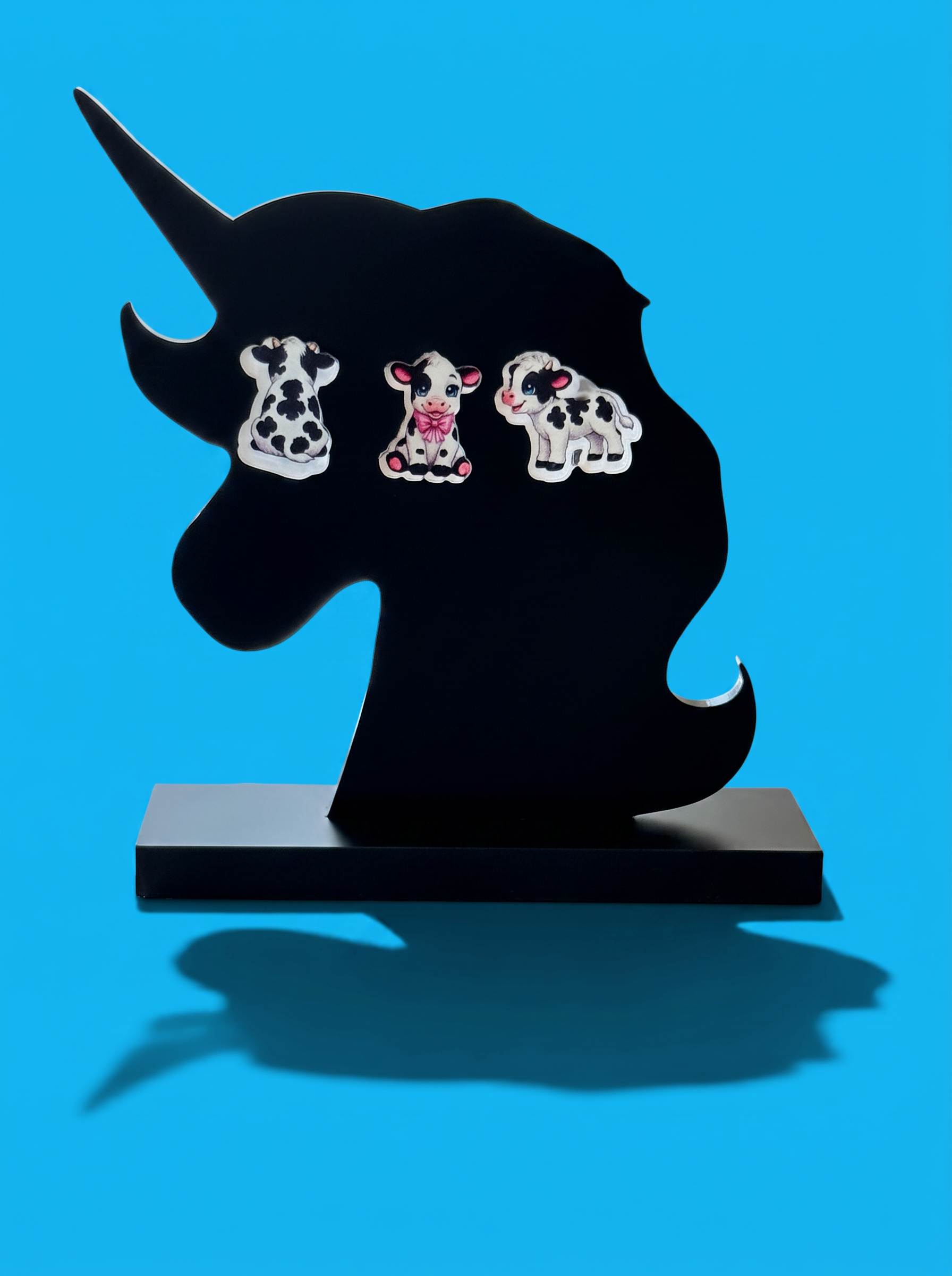 Three cow-shaped earrings displayed on a black unicorn head stand against a bright blue background.