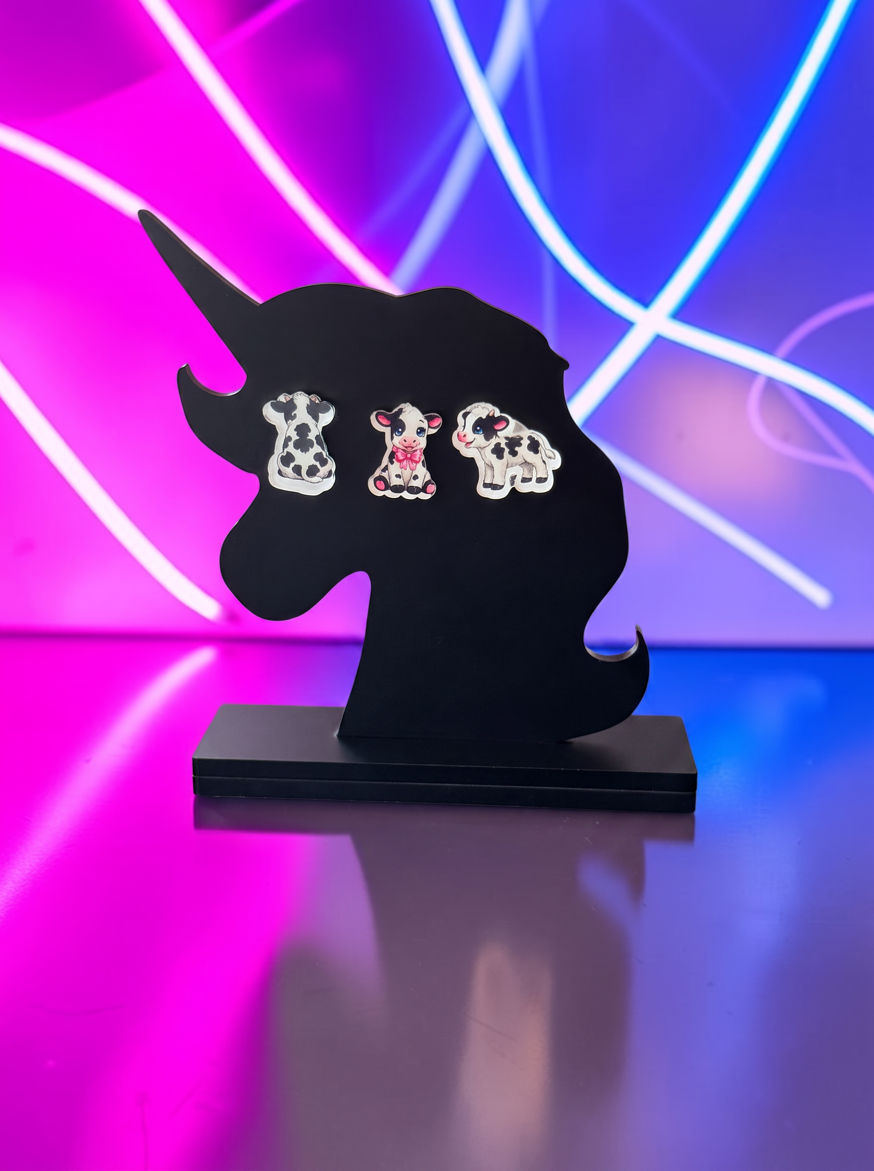Three cow-shaped stud earrings displayed on a black unicorn-shaped stand, set against a vibrant pink and blue neon light background.