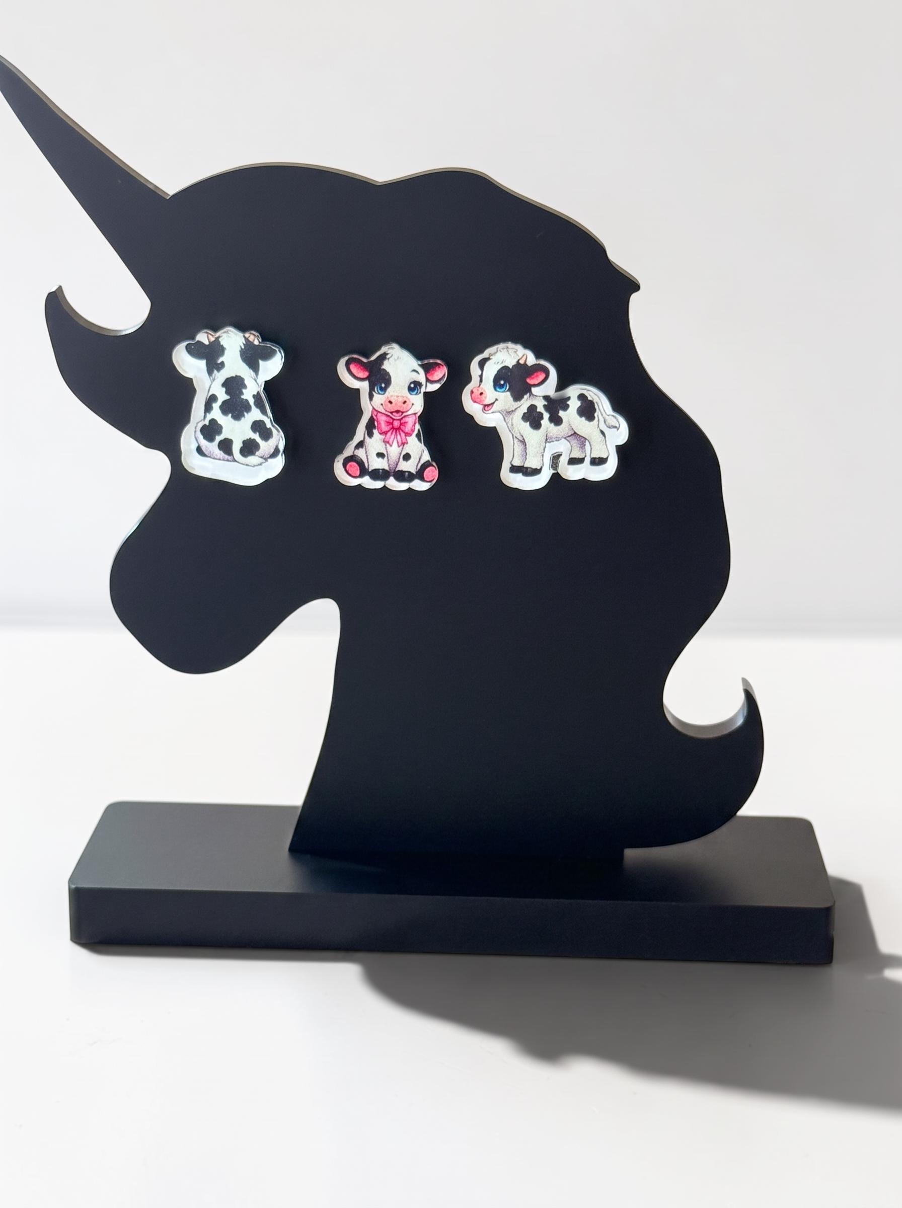 Three cow-shaped stud earrings displayed on a black unicorn-head stand against a plain light background.