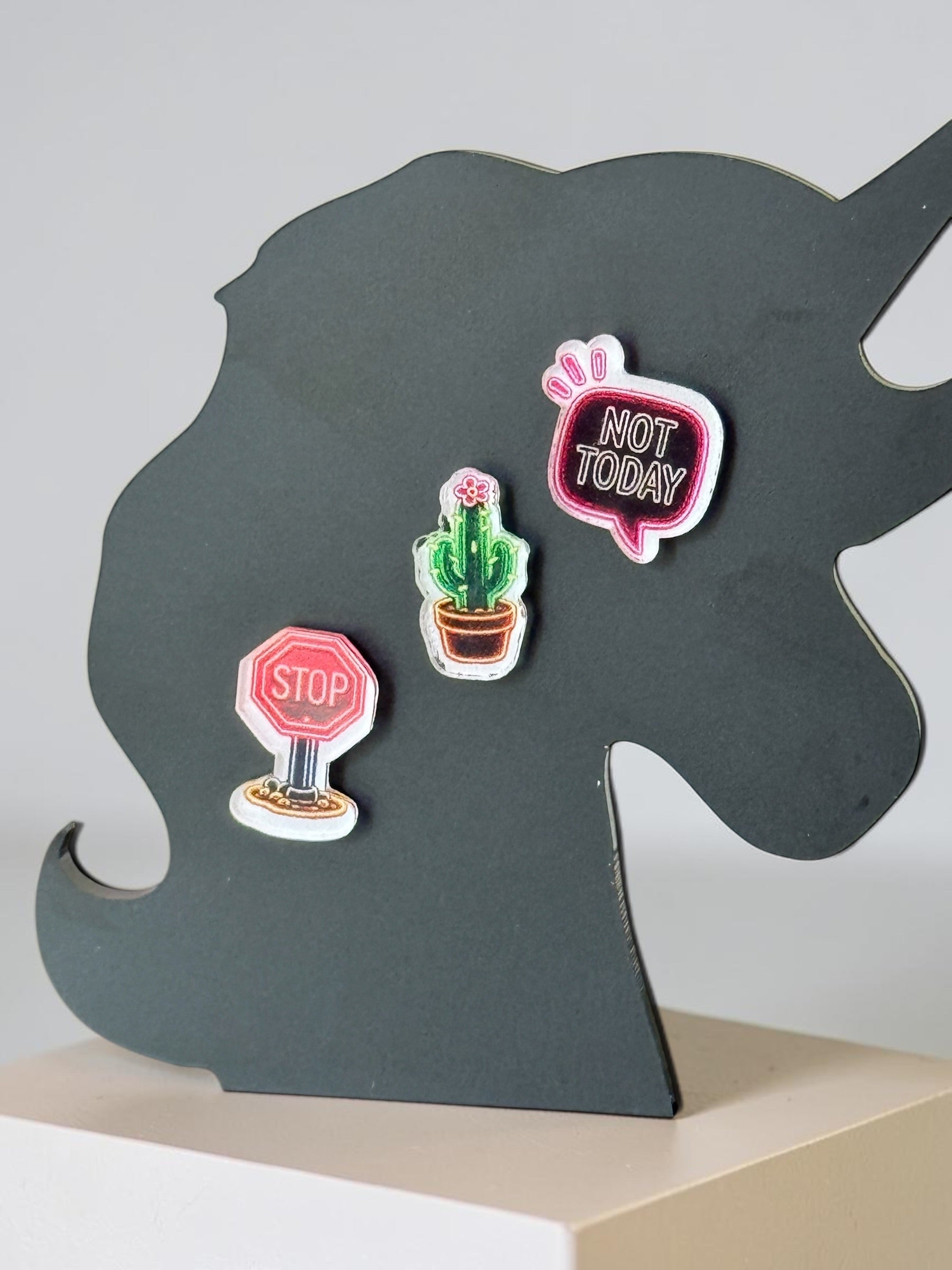 Three small decorative earrings displayed on a dark gray unicorn-shaped stand: a pink speech bubble sticker reading "NOT TODAY," a red stop sign sticker, and a green cactus in a pot sticker.