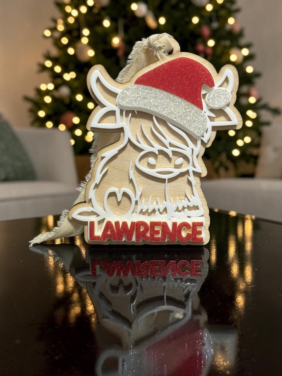 Personalized Highland cow Christmas ornament with a natural wood backer, white acrylic details, red glitter Santa hat and Lawrence name