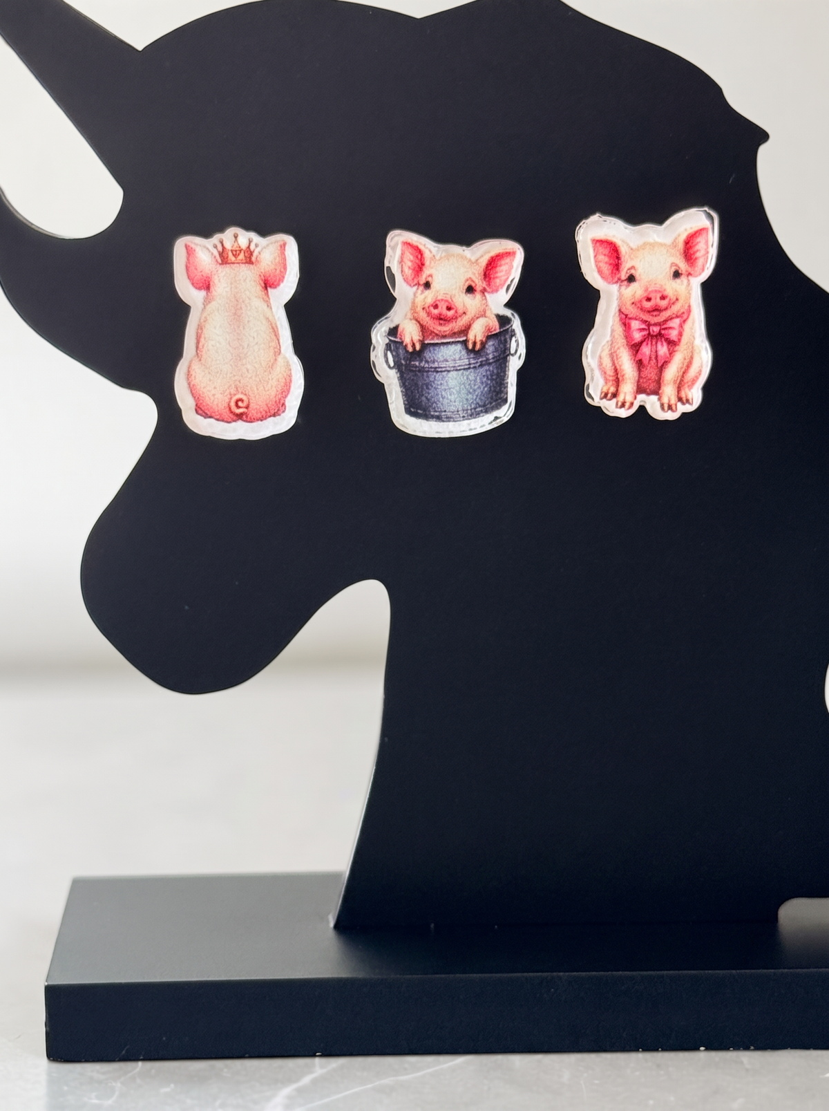 Three pig-themed earrings displayed on a black unicorn-shaped stand against a light gray background.