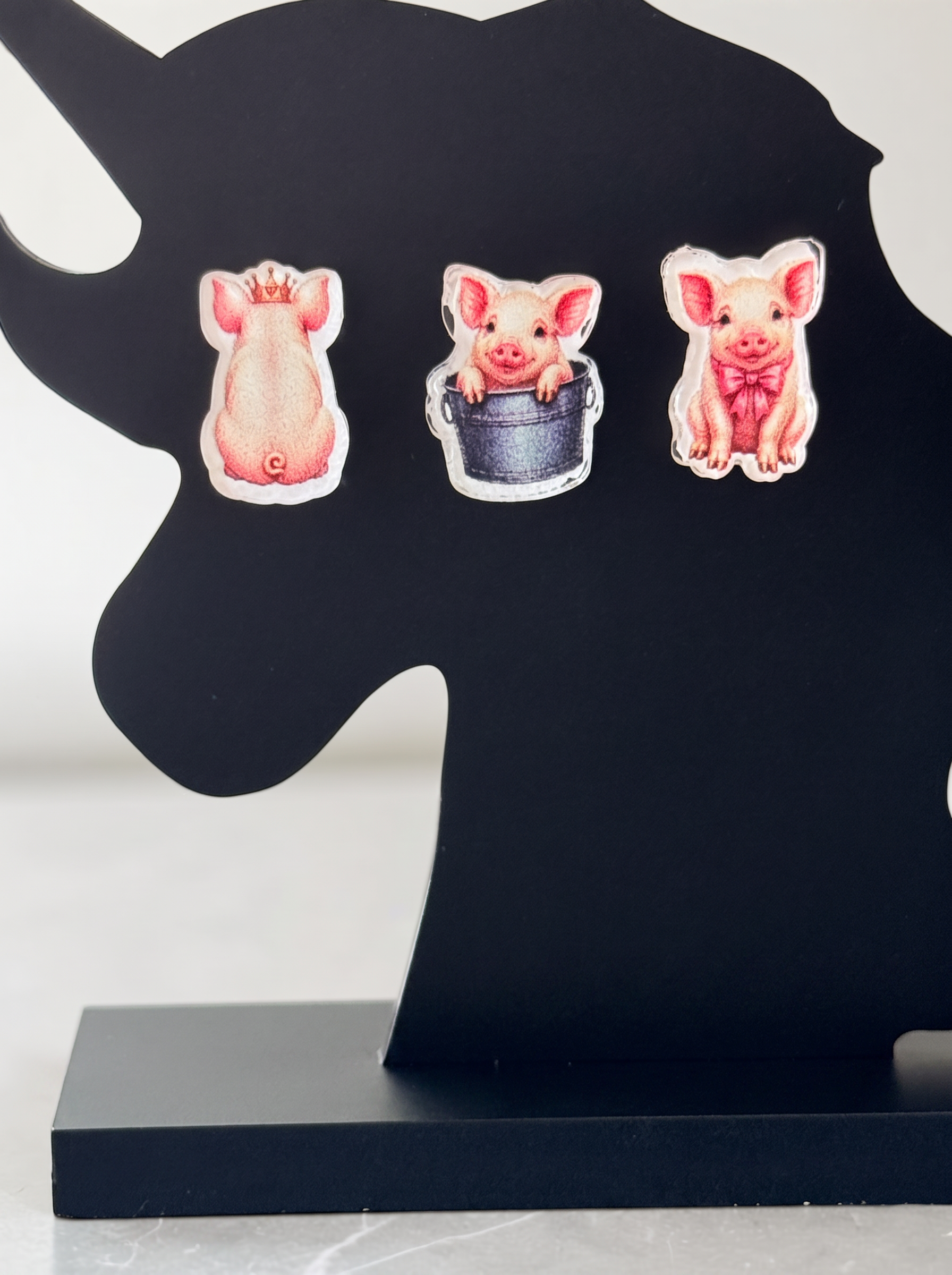 Three pig-themed earrings displayed on a black unicorn-shaped stand against a light gray background.