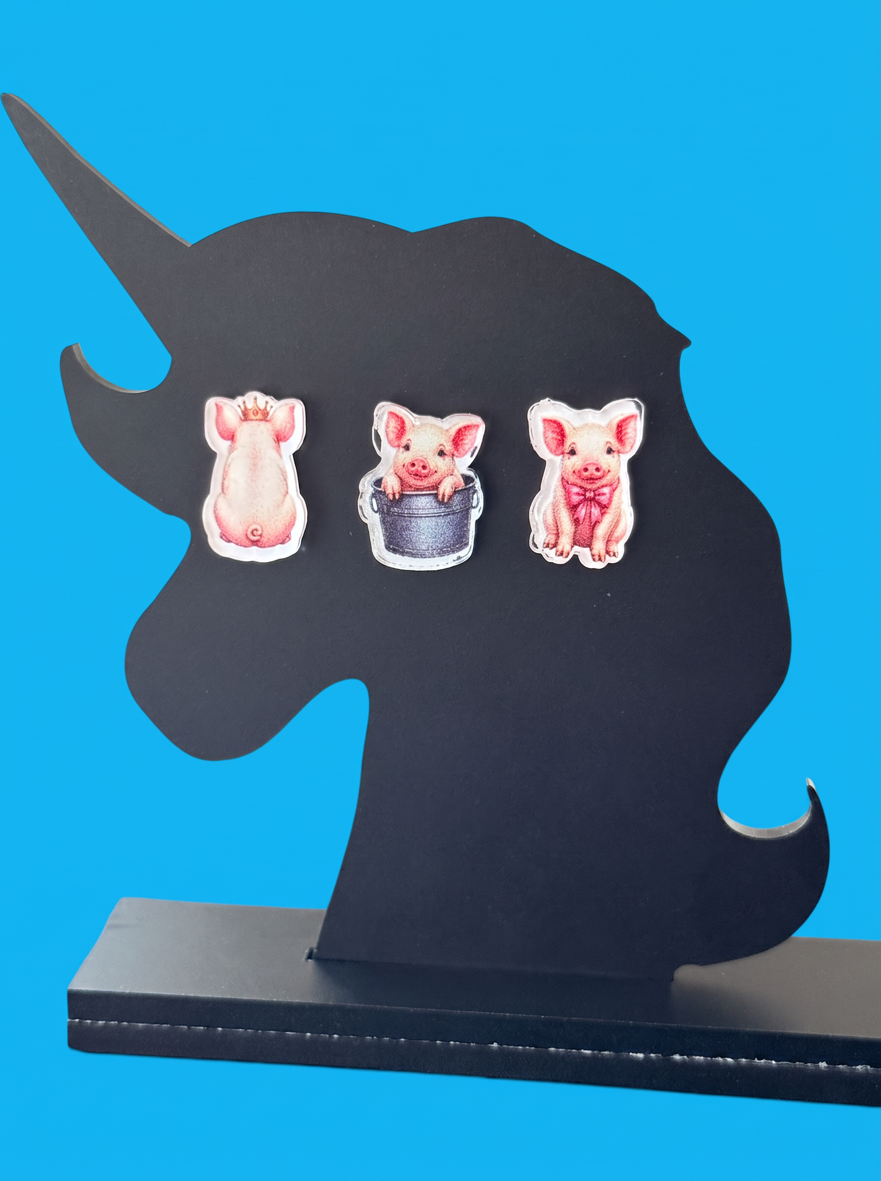 Three small pig-themed earrings displayed on a black unicorn-shaped stand against a bright blue background.