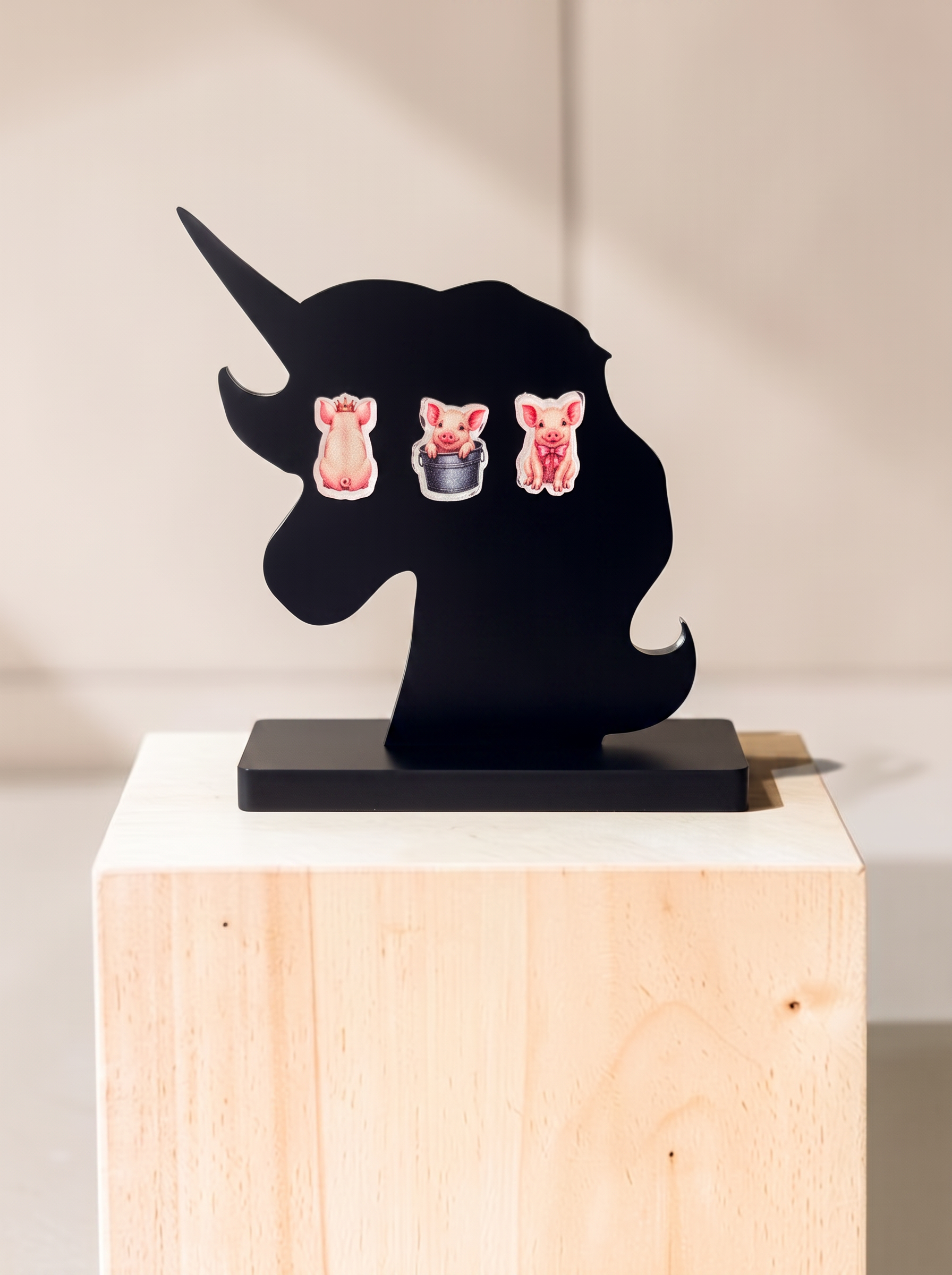 Three small animal stud earrings displayed on a black unicorn-shaped stand, which sits on a light wood block against a warm beige background.