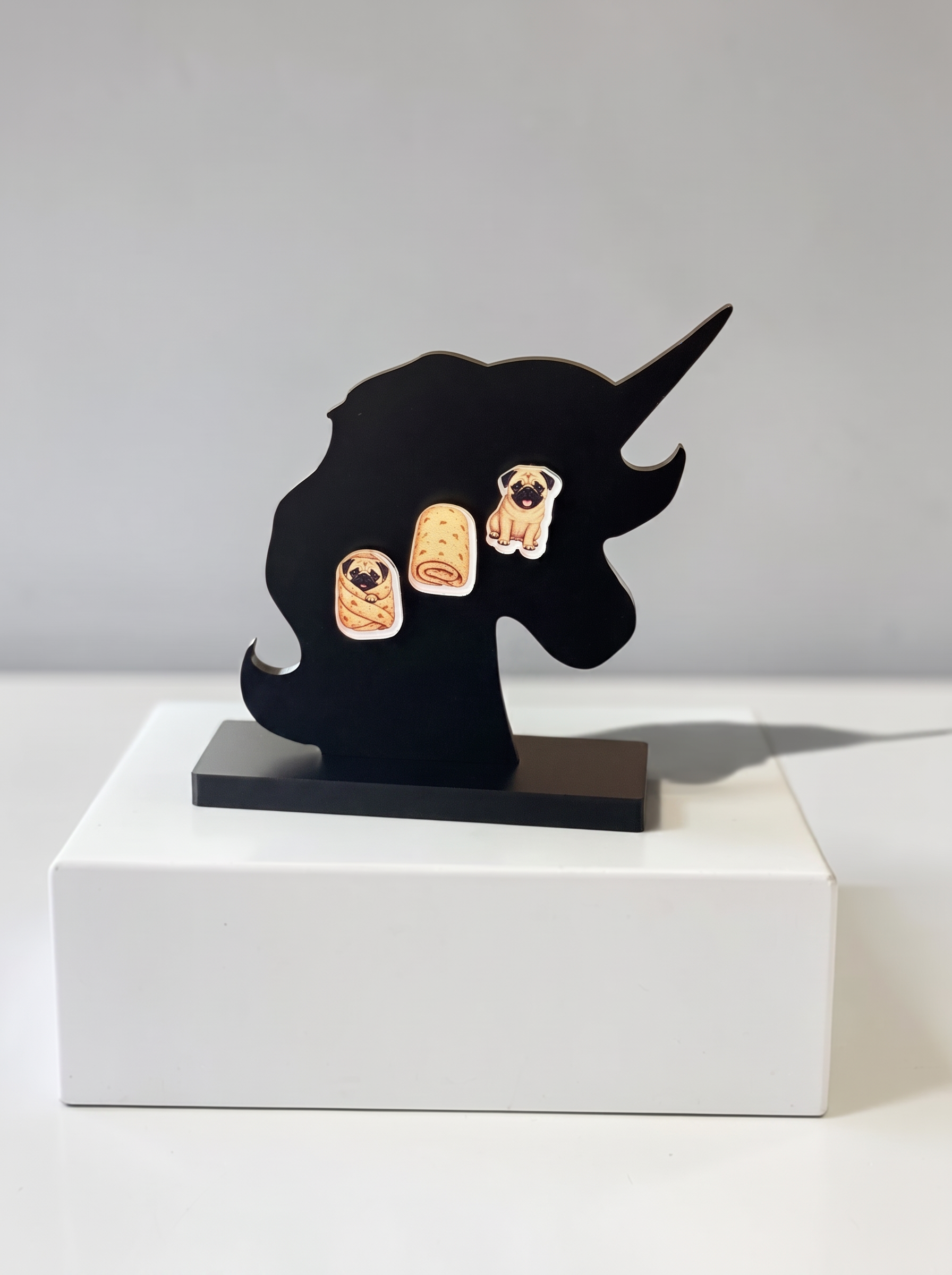 Three pug-themed earrings. The earrings depict a pug  wrapped in a burrito blanket, an image of the burrito blanket, and sitting proudly.shown on a white rectangular pedestal against a light gray background.
