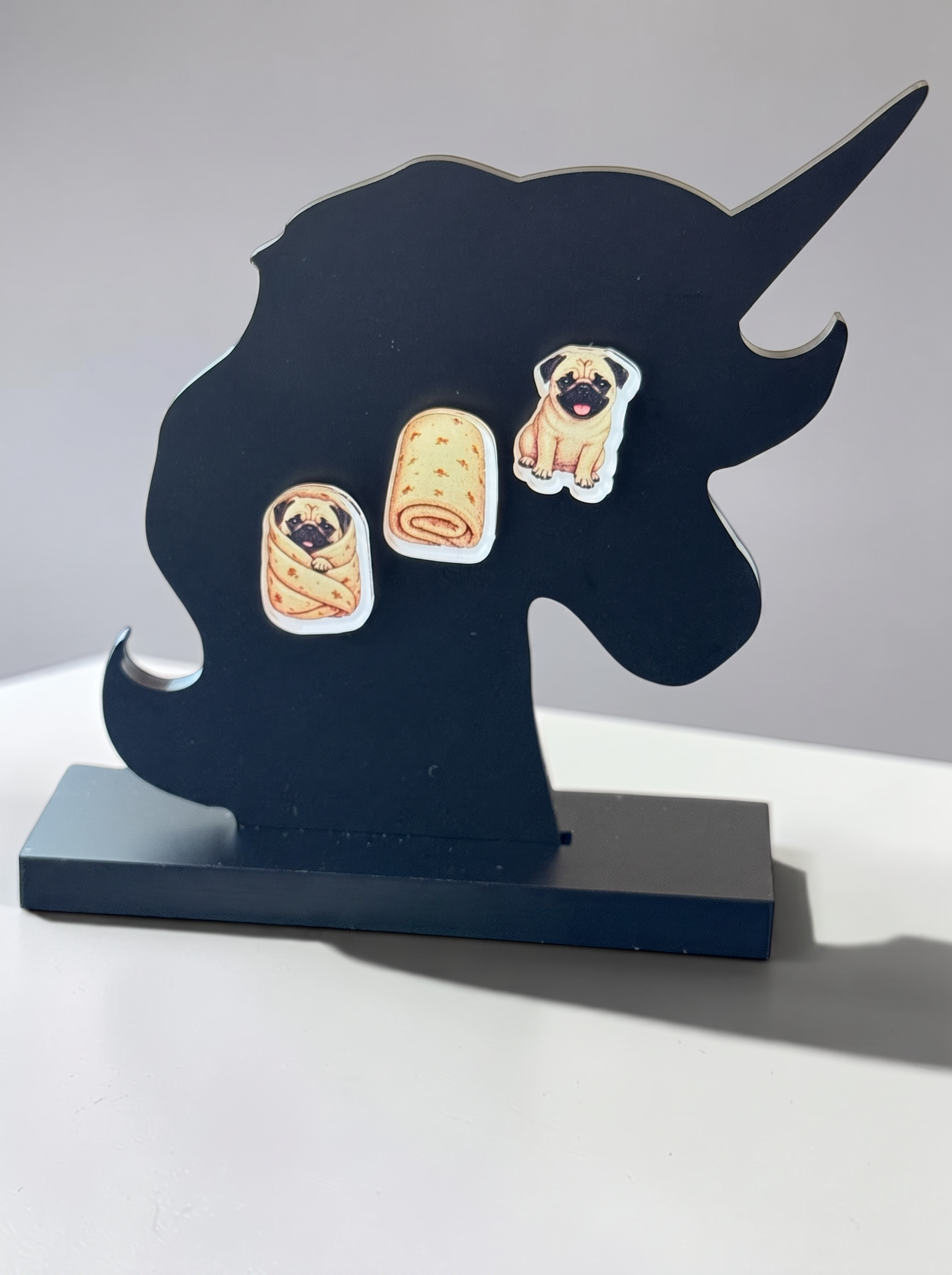 A black unicorn-shaped decorative stand with a rectangular base, featuring three small illustrated pug earrings, photographed on a light gray background.