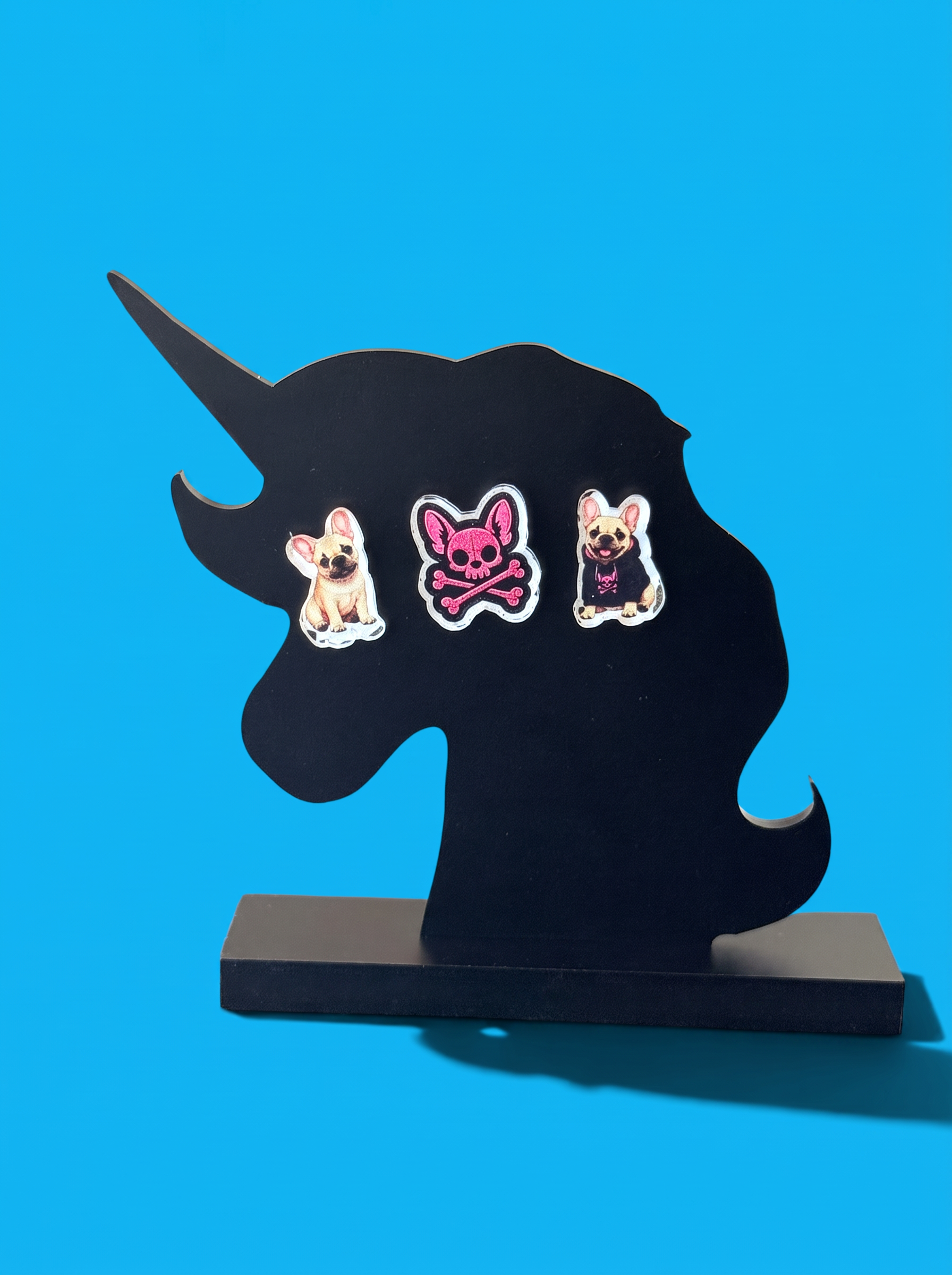 Three decorative earrings featuring French bulldogs and a pink skull-and-crossbones motif, displayed on a black unicorn-shaped stand against a bright blue background.