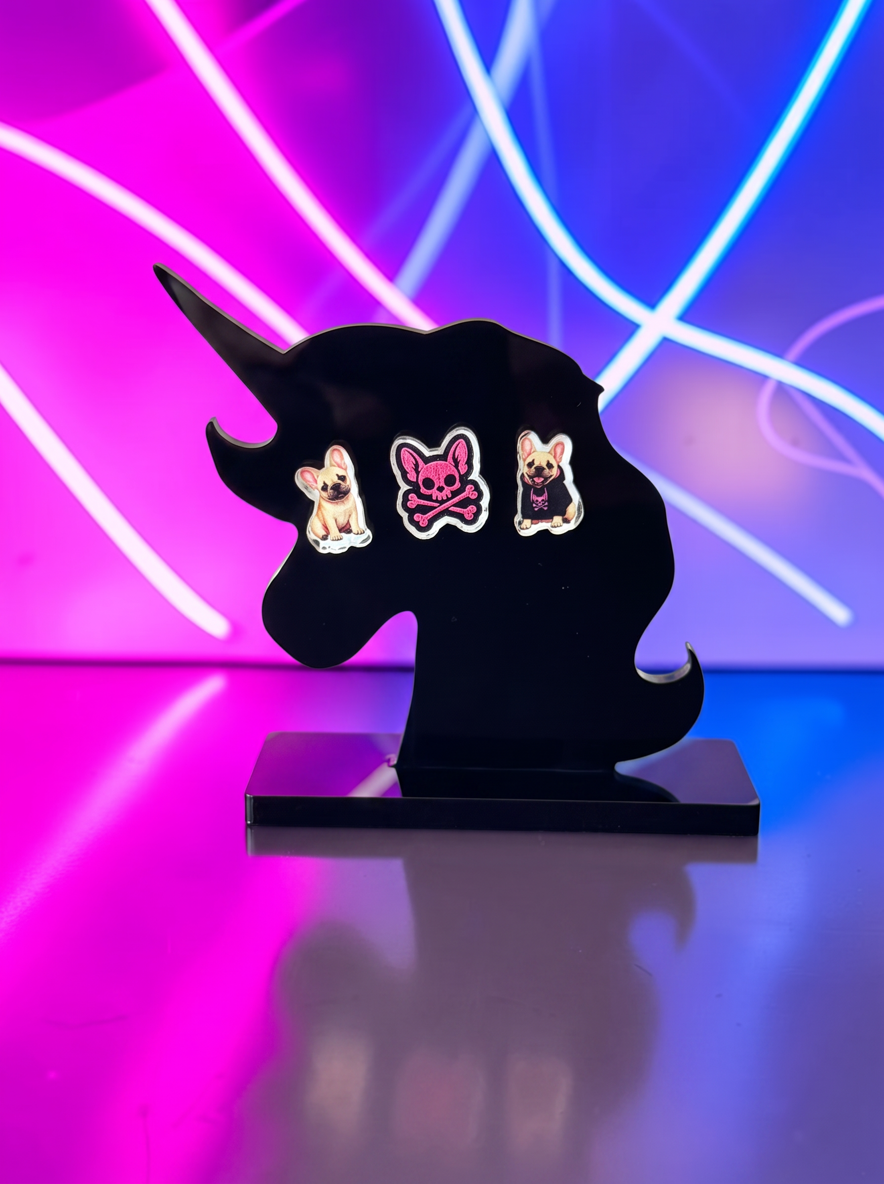 Three small dog-themed earrings displayed on a black unicorn-shaped stand against a neon pink and blue light background.