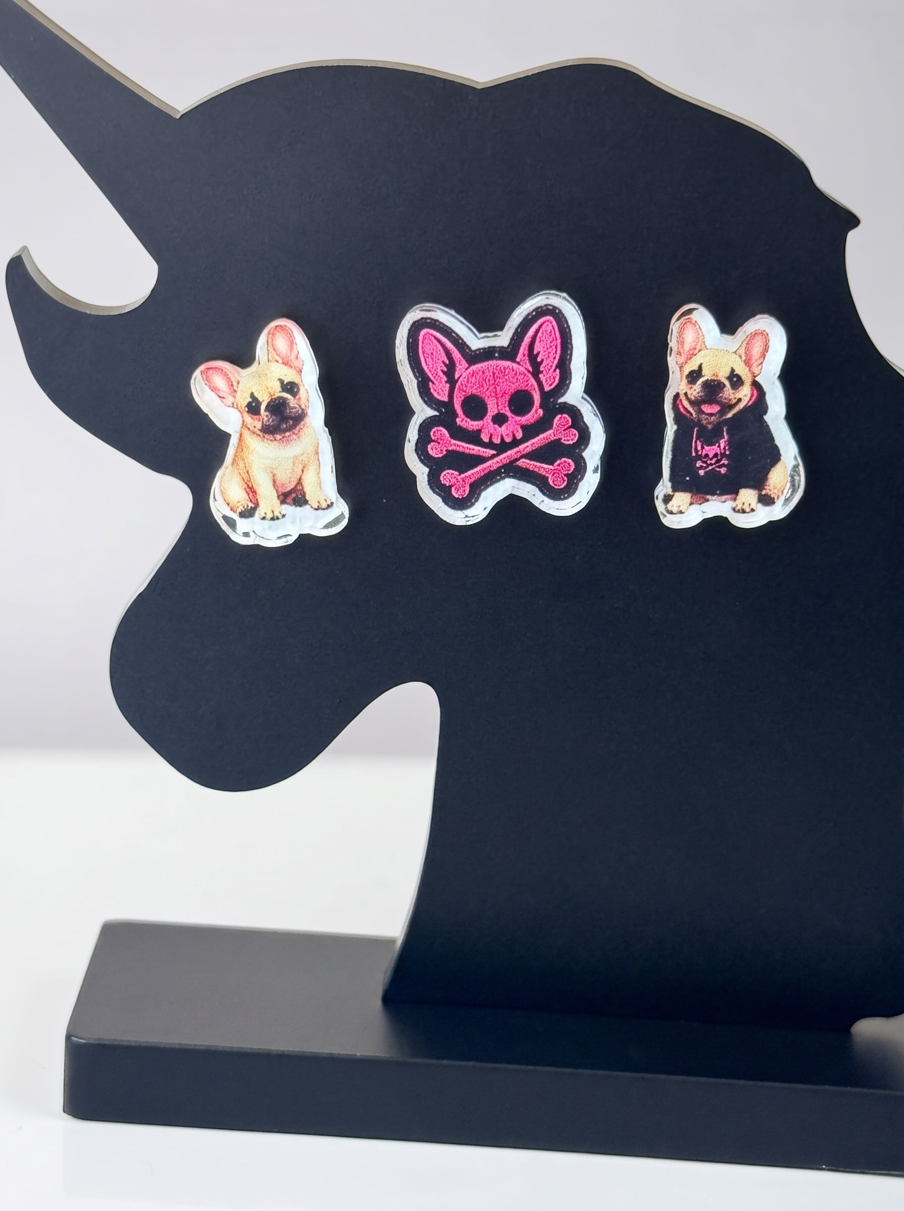 Three decorative stickers displayed on a black unicorn-shaped stand. The earrings feature a tan French Bulldog, a pink skull and crossbones motif, and another tan French Bulldog wearing a black hoodie.