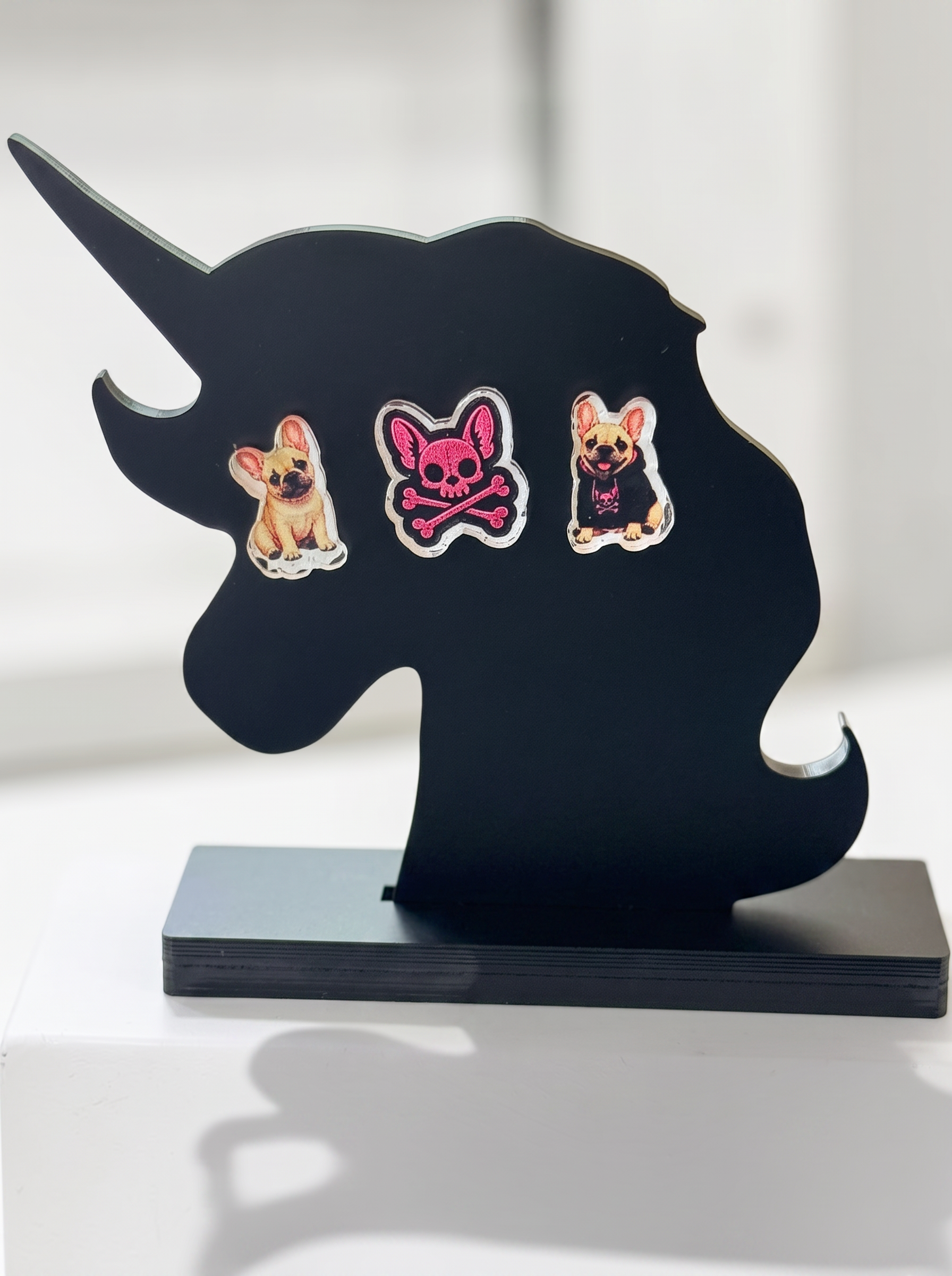 A black unicorn-shaped display stand with a rectangular base, featuring three decorative earrings. The earrings include a tan French Bulldog, a pink skull-and-crossbones dog with white details, and a pink French Bulldog wearing a black shirt.