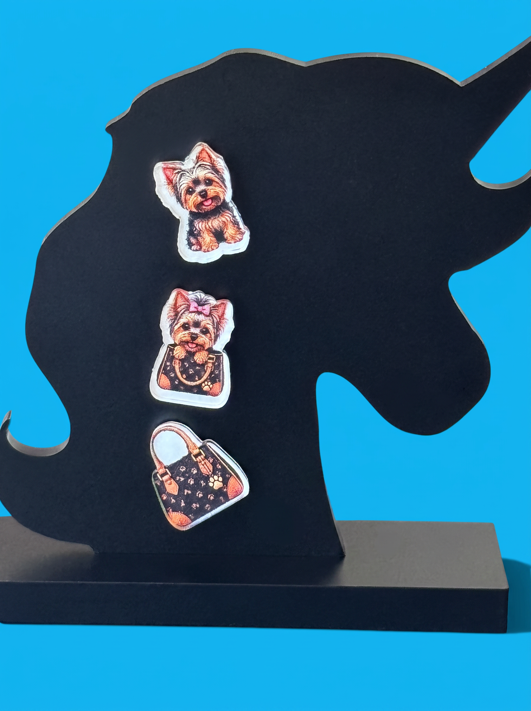 Three decorative yorkie-themed earrings displayed on a black unicorn-shaped stand against a bright blue background.