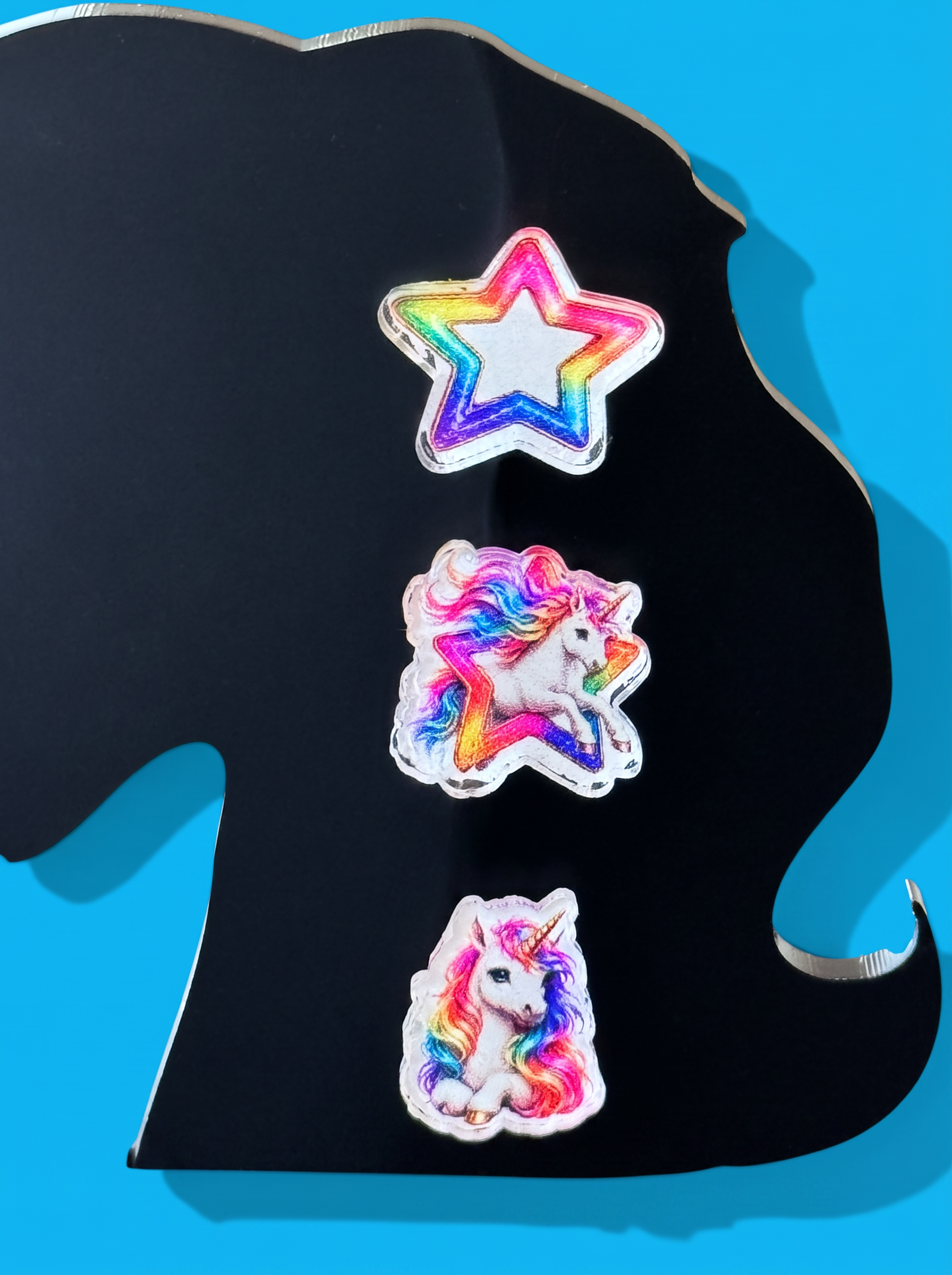 Three colorful unicorn-themed earrings displayed on a black stand against a bright blue background. The earrings include a rainbow star, a rainbow unicorn, and a white unicorn with a horn and rainbow mane.