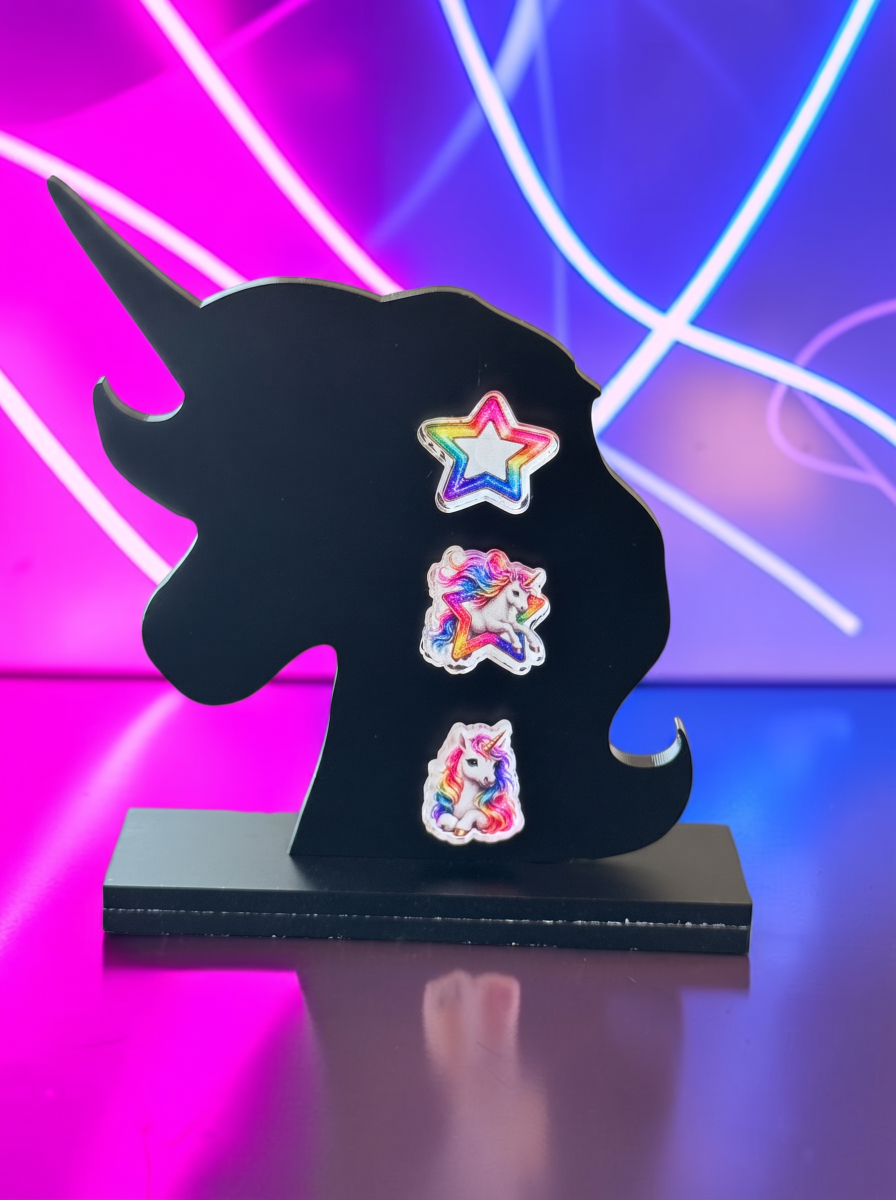Three colorful unicorn-themed earrings displayed on a black stand against a bright blue background. The earrings include a rainbow star, a rainbow unicorn, and a white unicorn with a horn and rainbow mane, set against a neon pink and blue light background.