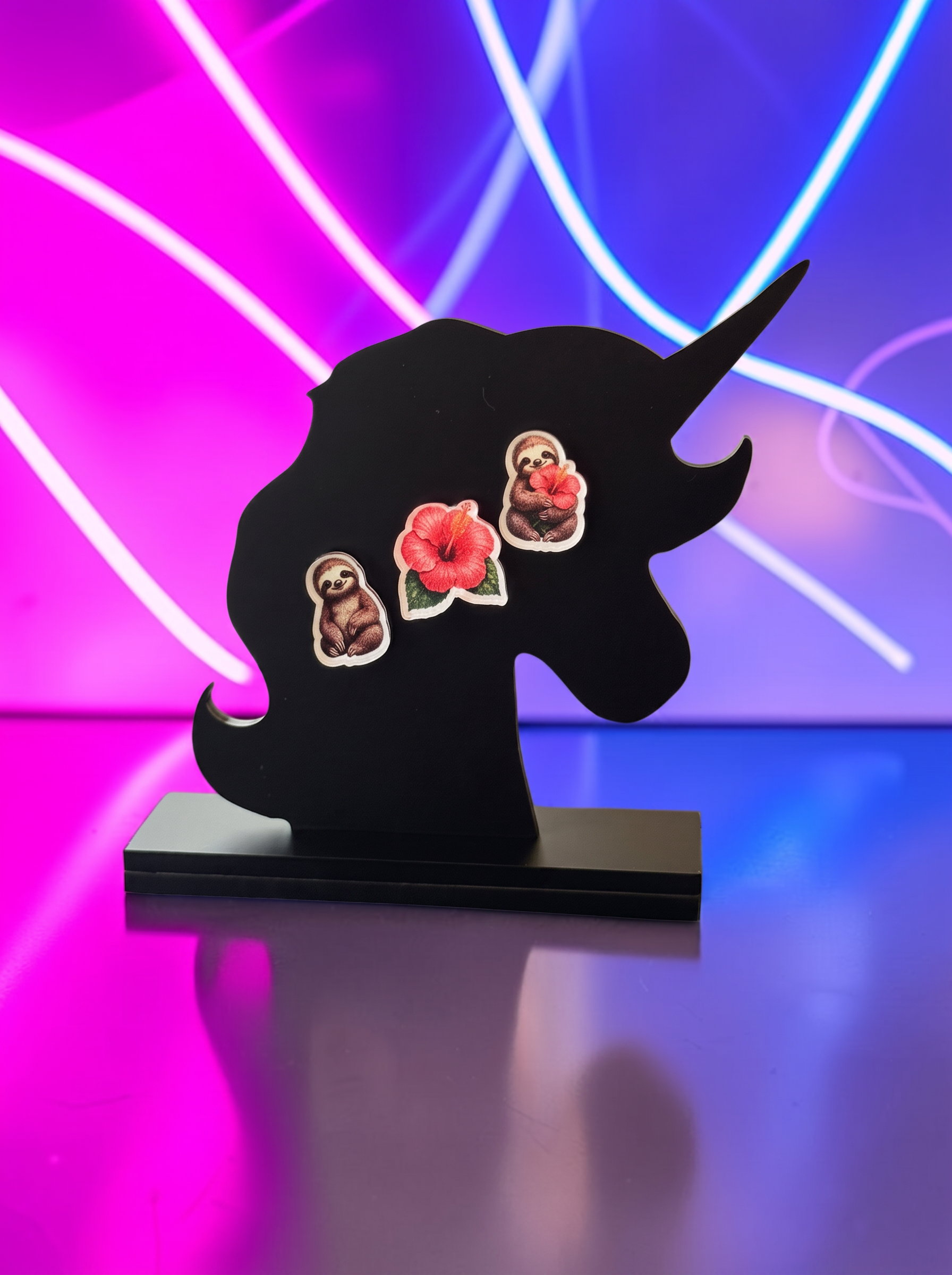A black unicorn-shaped decorative plaque or stand with a rectangular base, featuring three small decorative earrings: two sloths and a hibiscus flower. The background features vibrant pink and blue neon light tubes.